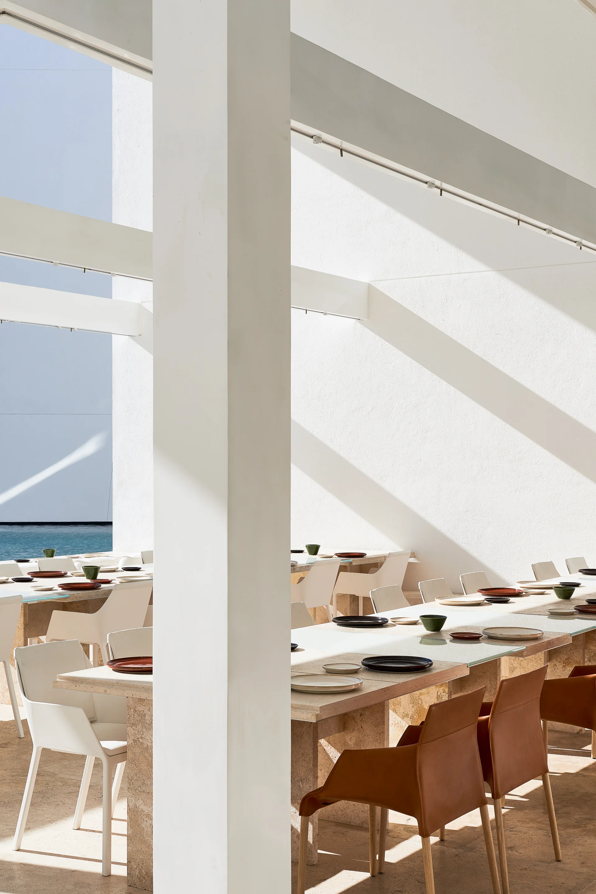 An open-air dining patio features stone-topped tables, white and leather chairs, and large white stucco columns under structural beams.