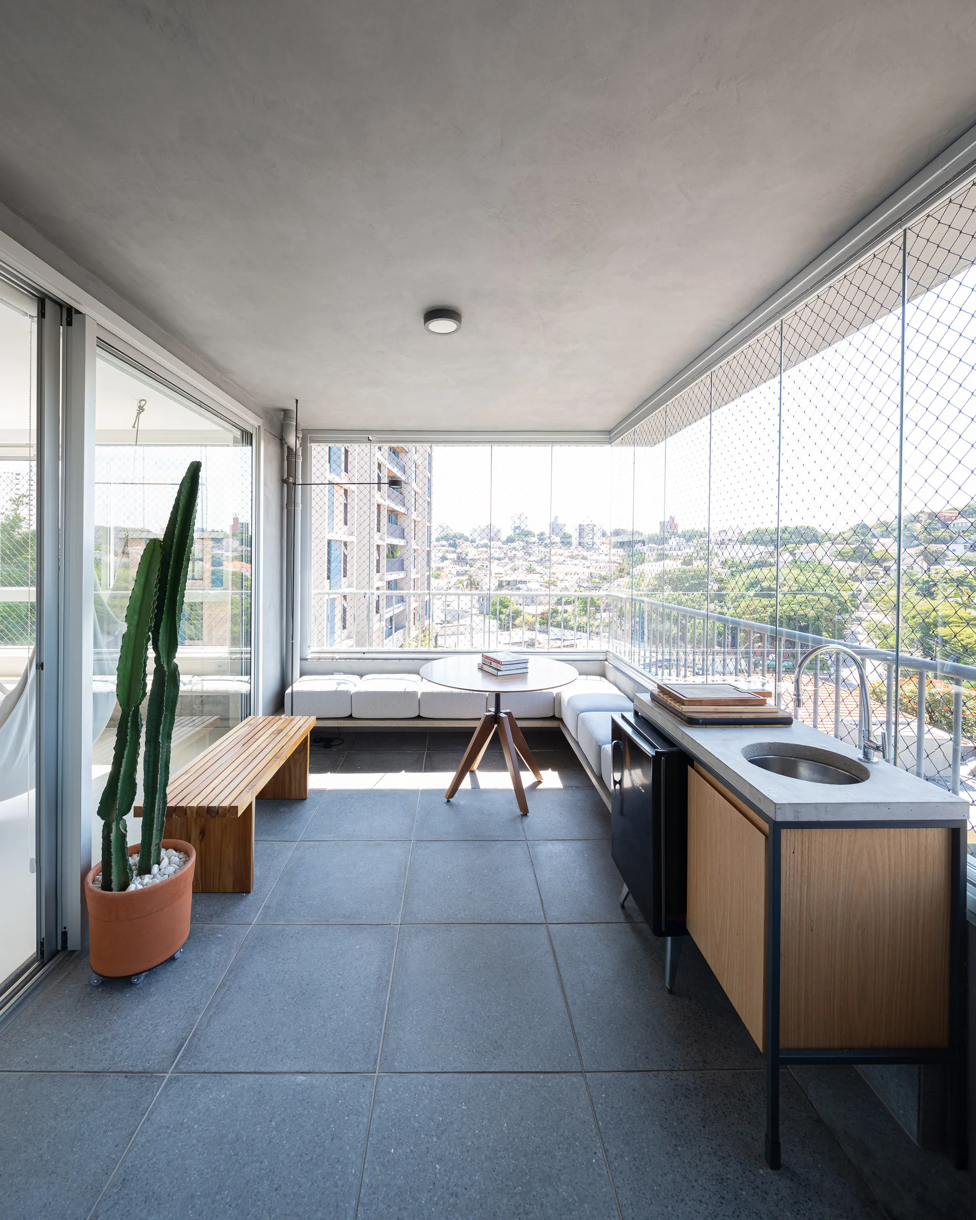 An enclosed balcony with a built-in L-shaped bench, a round wooden dining table, and a small outdoor kitchen with a concrete countertop.