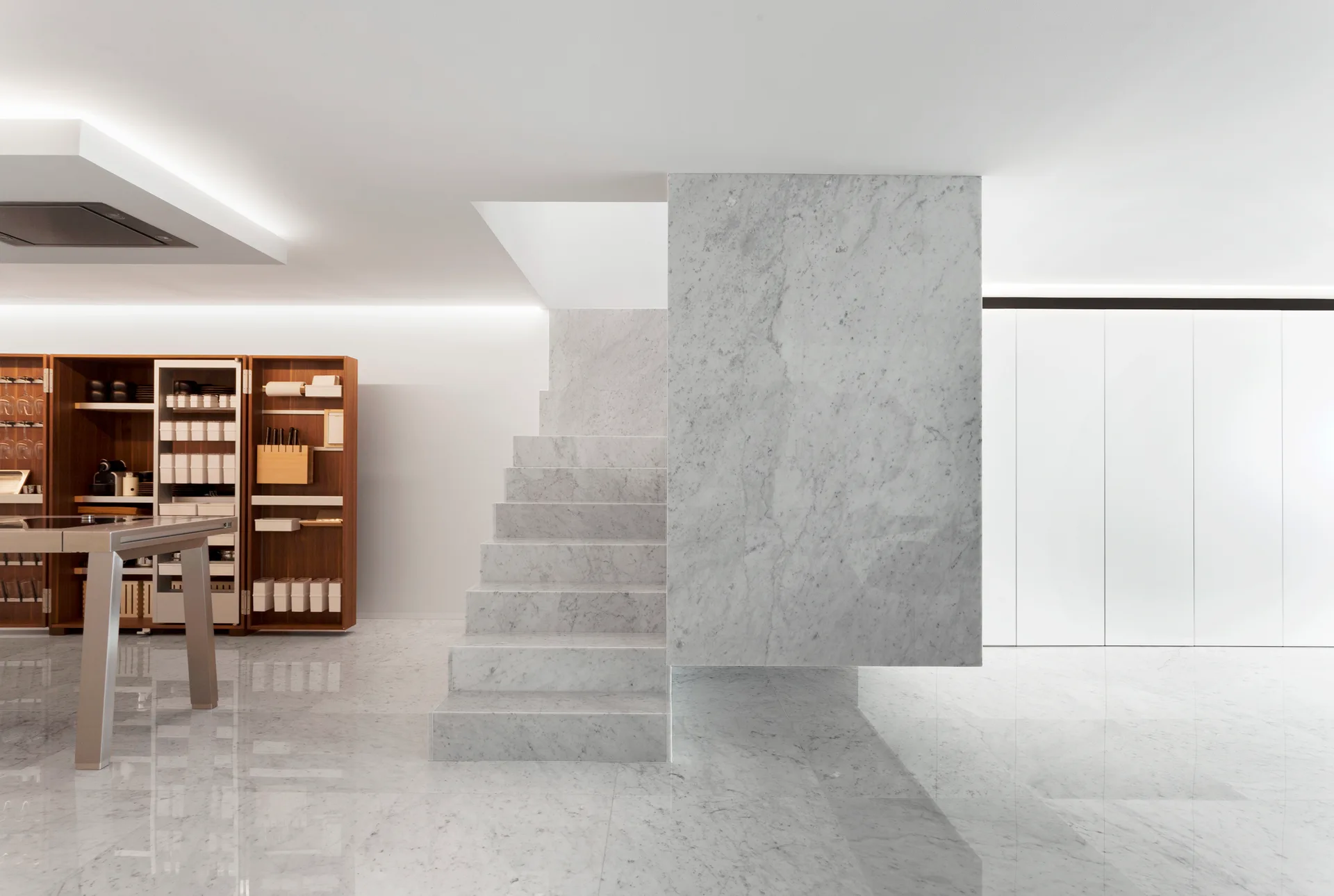 A minimalist modern kitchen featuring a sculptural grey-veined white marble staircase, polished marble flooring, and a tall wood pantry unit.
