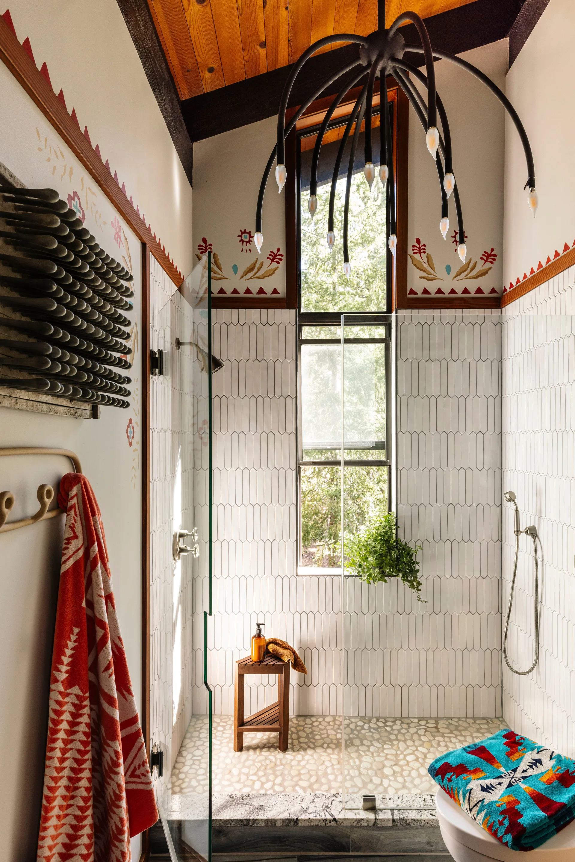 An eclectic bathroom features a glass-enclosed shower with vertical white tile, pebble flooring, a wood ceiling, and a black sculptural chandelier.