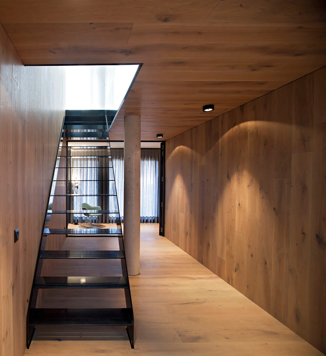 A minimalist hallway features wood paneled walls and ceilings, a concrete column, and an open-tread black metal staircase.