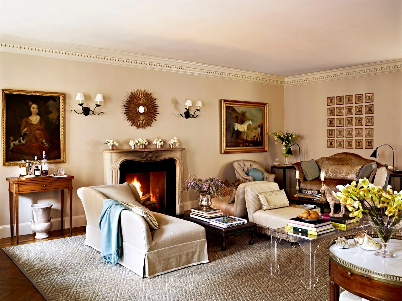 A traditional living room features beige walls, a stone fireplace with a burning fire, upholstered armchairs, and framed artwork on the walls.
