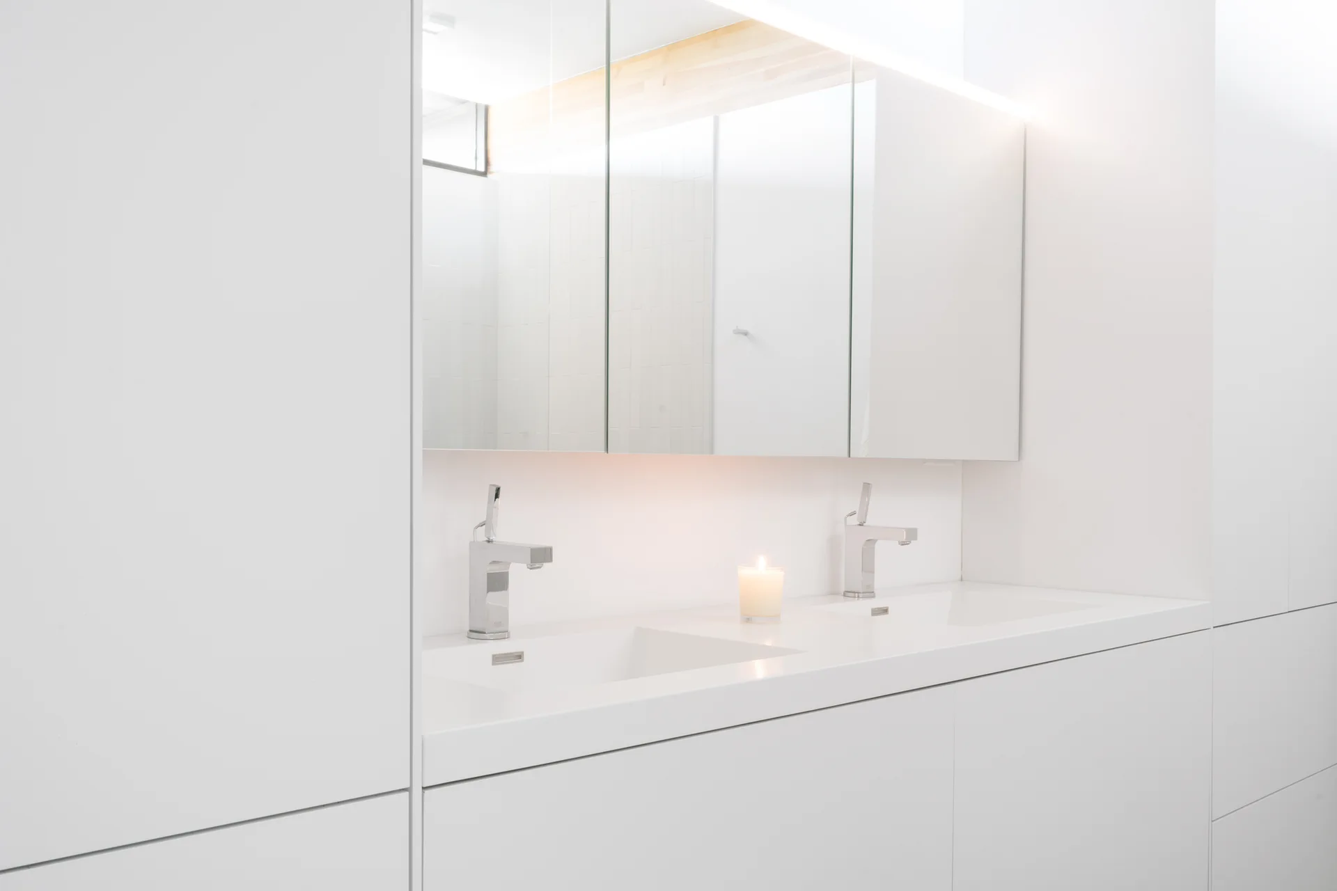 A minimalist white double bathroom vanity features chrome faucets under a mirrored medicine cabinet with warm under-cabinet lighting.