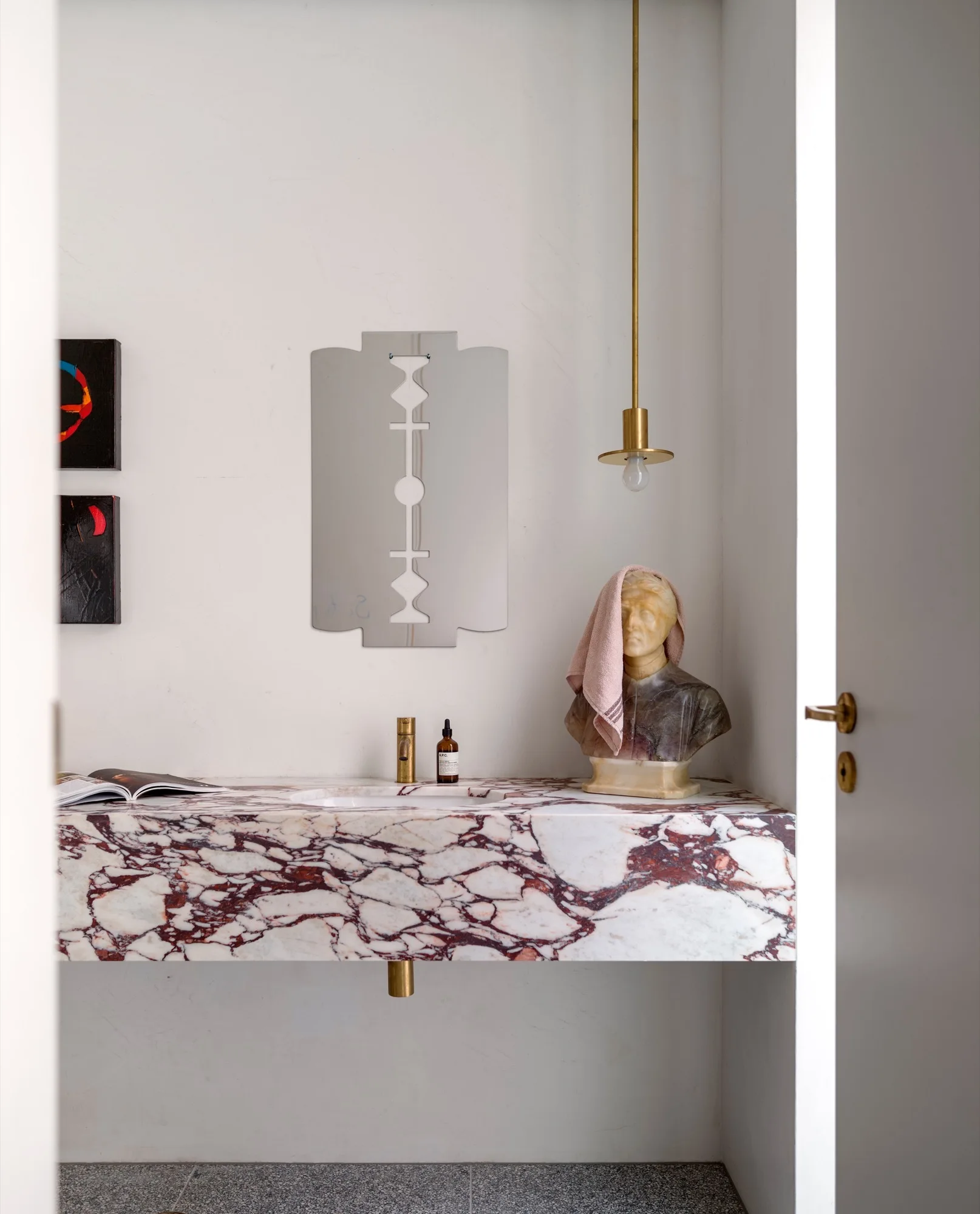 A floating purple-veined white marble bathroom vanity with a brass faucet, a razor-blade-shaped mirror, and a classic bust sculpture.