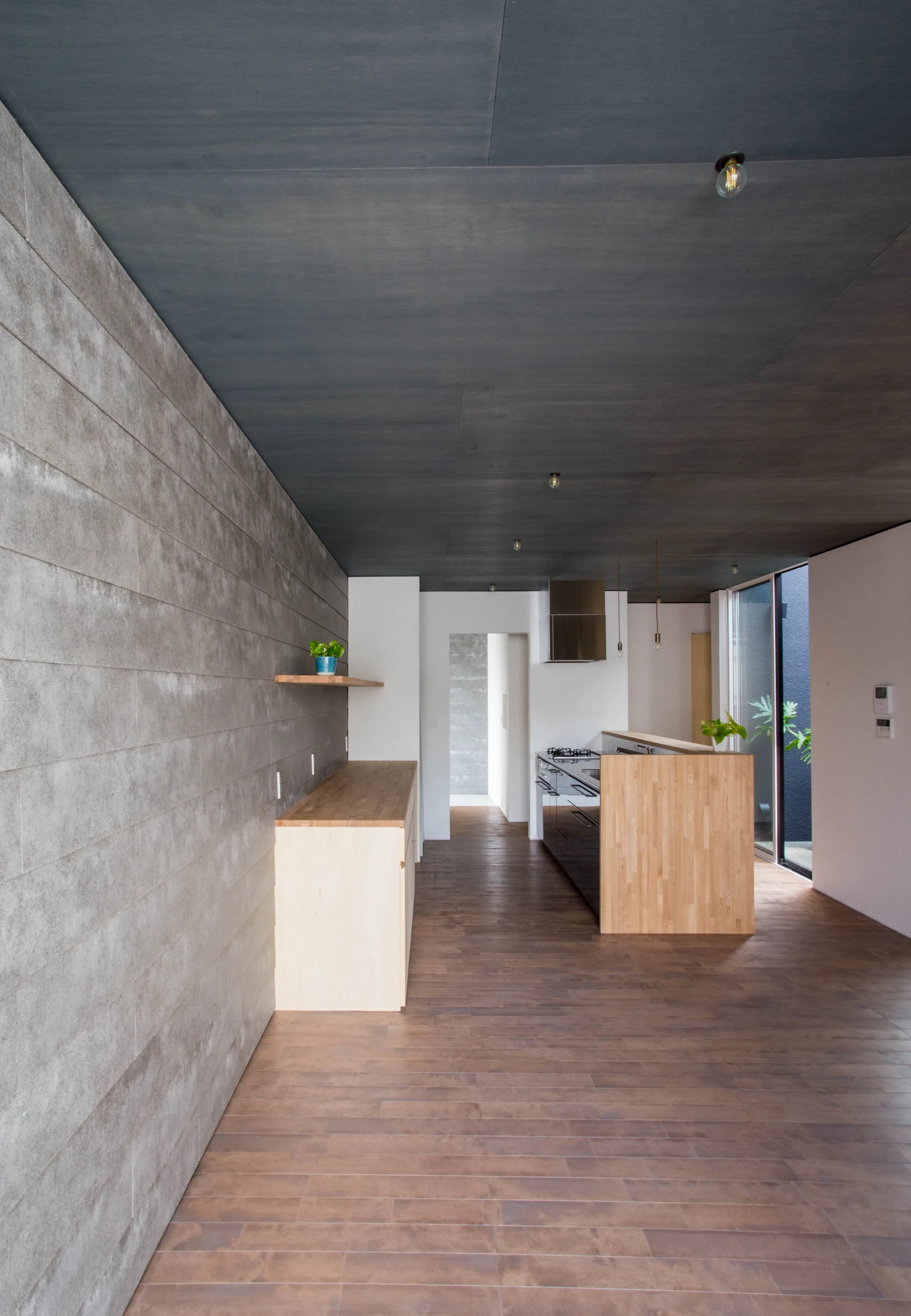 A minimalist kitchen features horizontal grey concrete wall panels, a light wood counter, a dark grey ceiling, and medium-toned wood plank flooring.