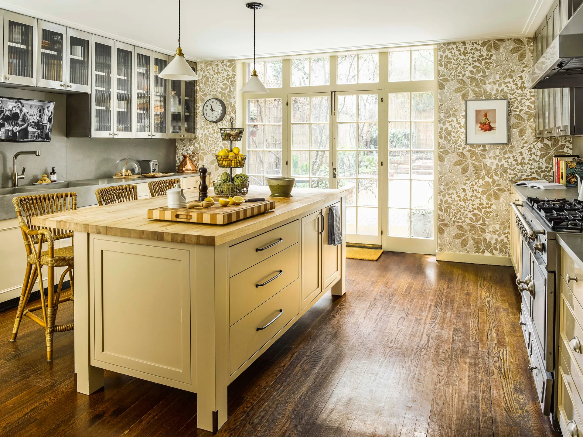 A traditional kitchen features a cream island with a butcher block top, botanical wallpaper, wicker stools, and multi-pane glass French doors.