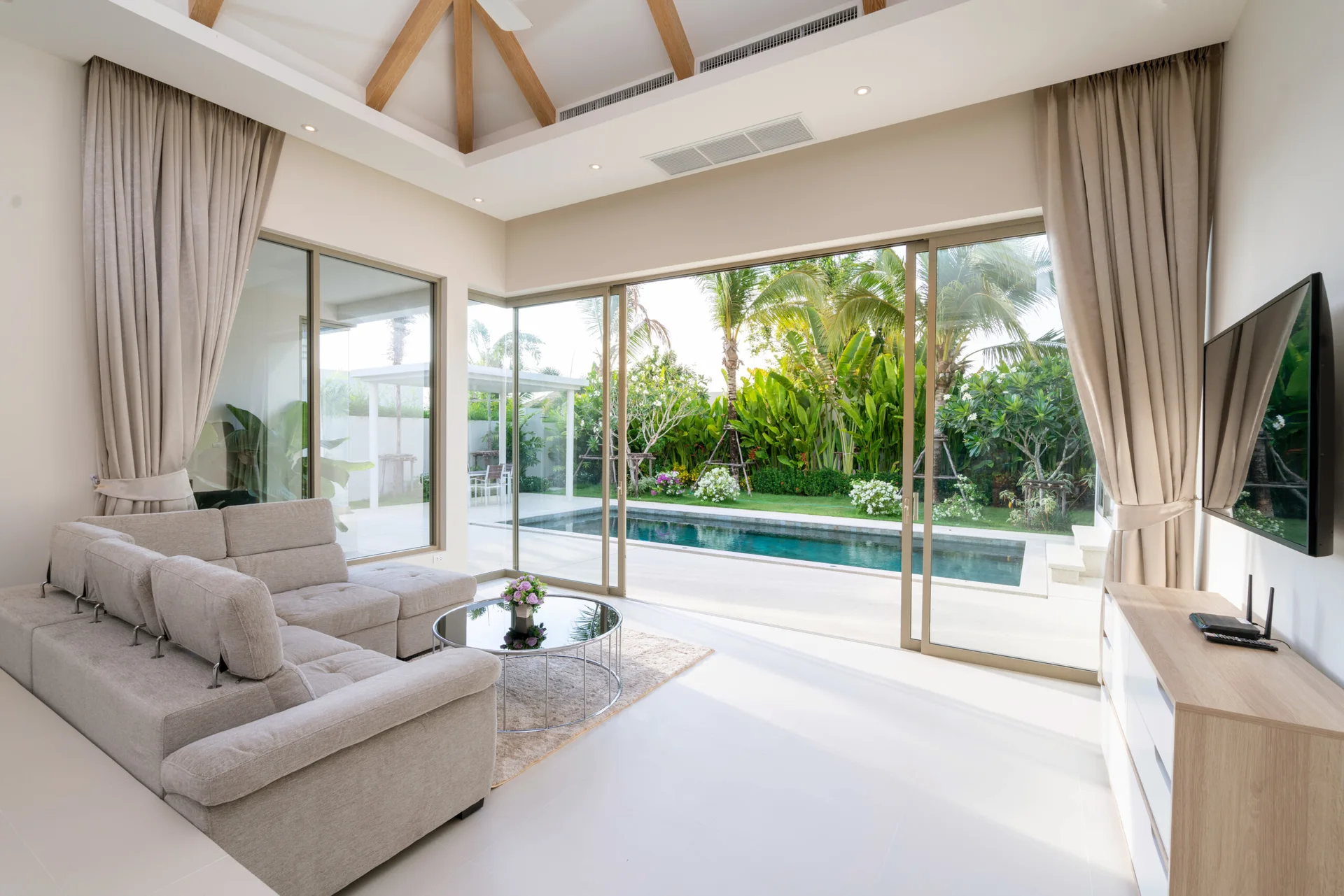 A bright living room with a beige sectional sofa, a round glass coffee table, and large sliding doors opening to an outdoor swimming pool.