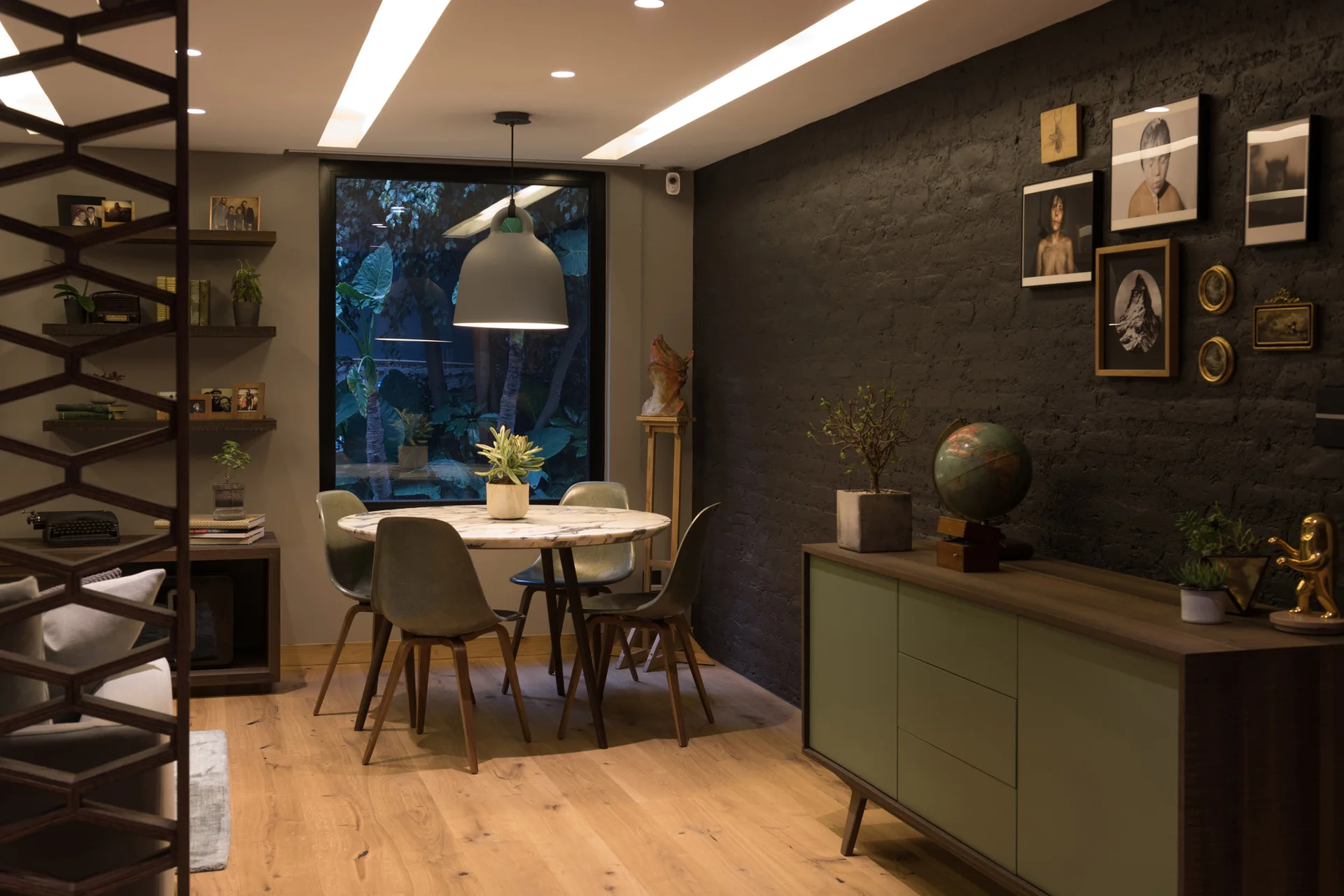A modern dining room features a round marble-topped table and green chairs set against a black brick wall with framed art and a green sideboard.