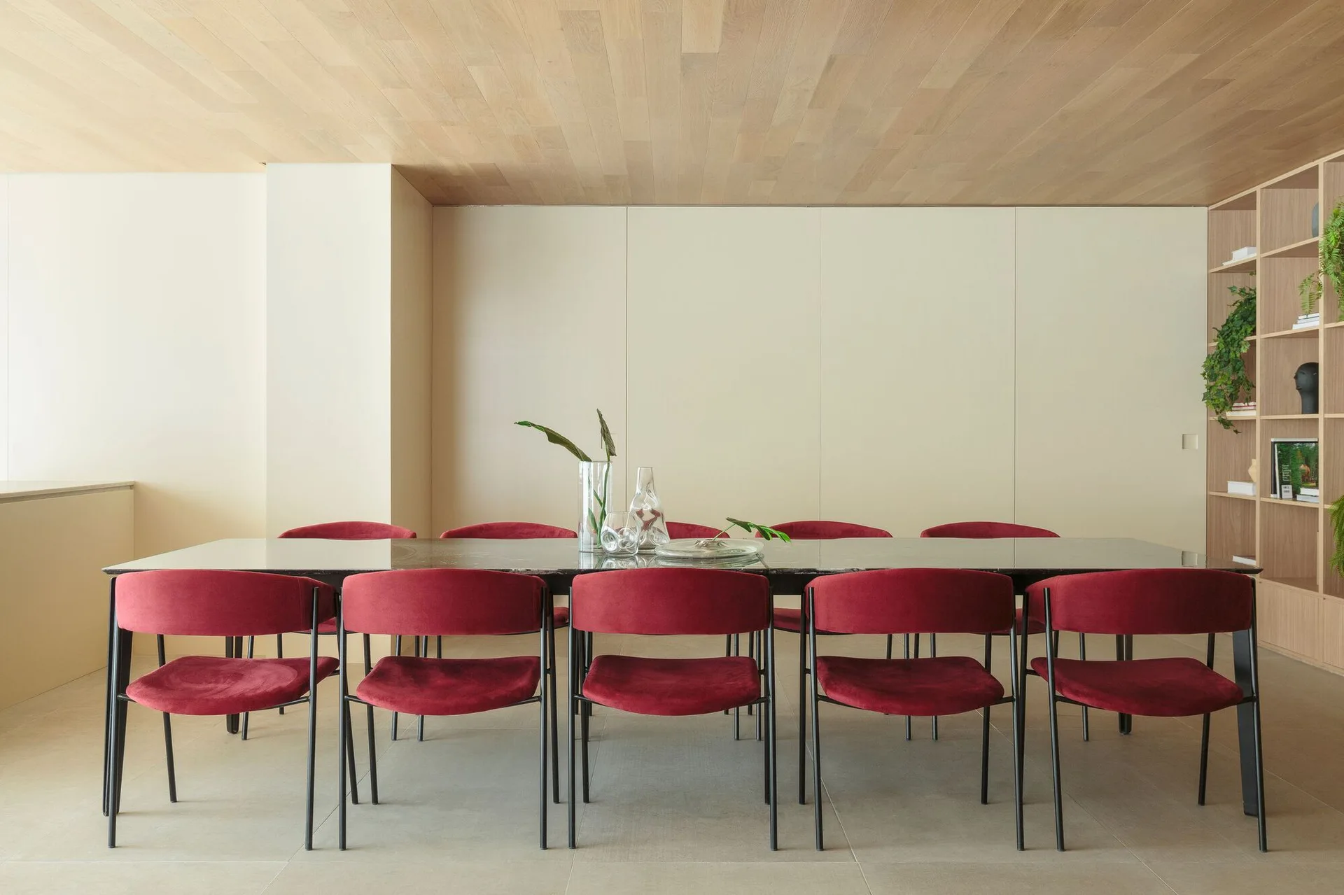 A minimalist dining room features a dark rectangular table surrounded by red velvet chairs under a light wood paneled ceiling.