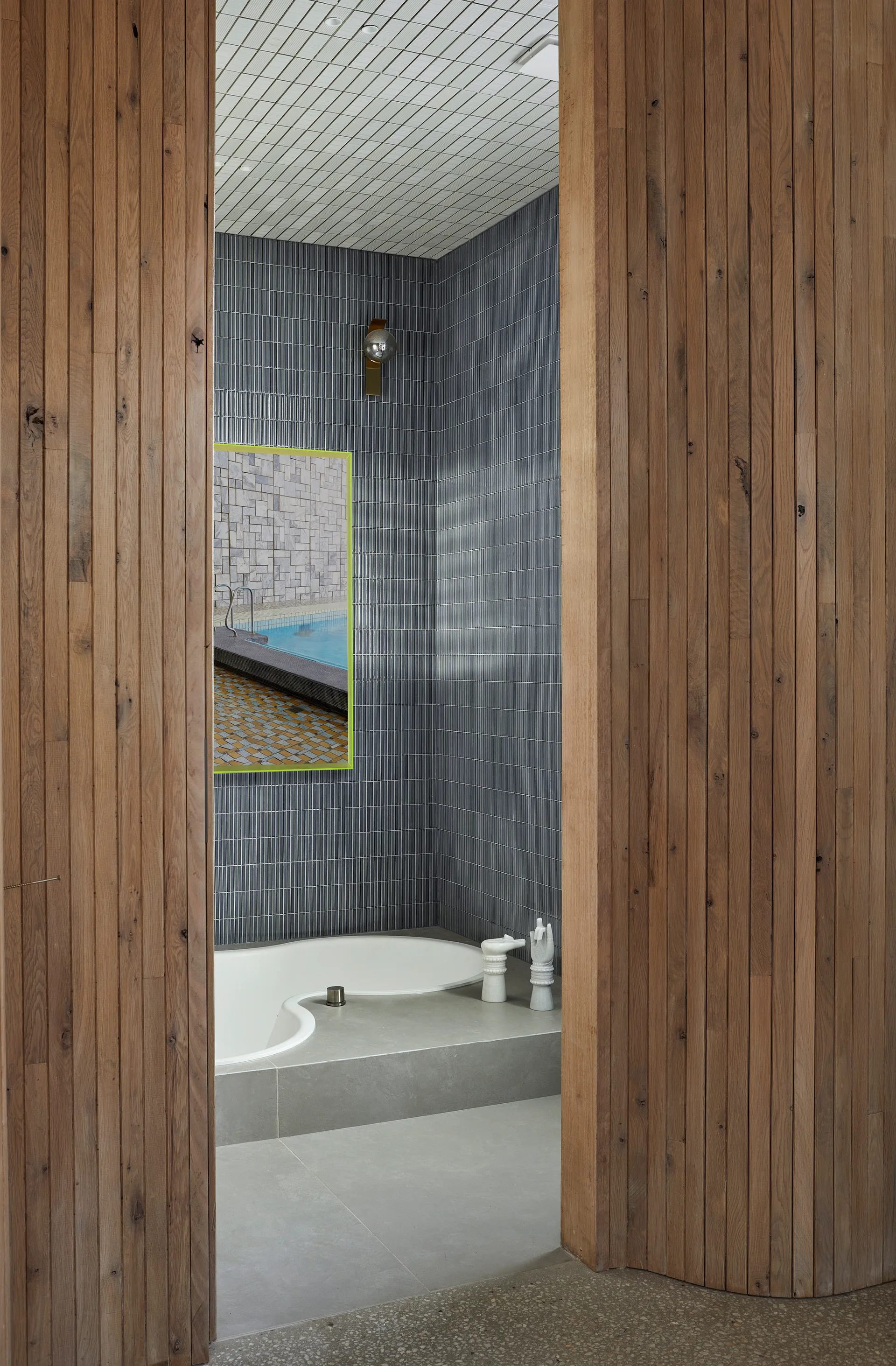A modern bathroom with a curved white bathtub, dark grey vertical tiled walls, and a neon green framed art print seen through a wood slat doorway.