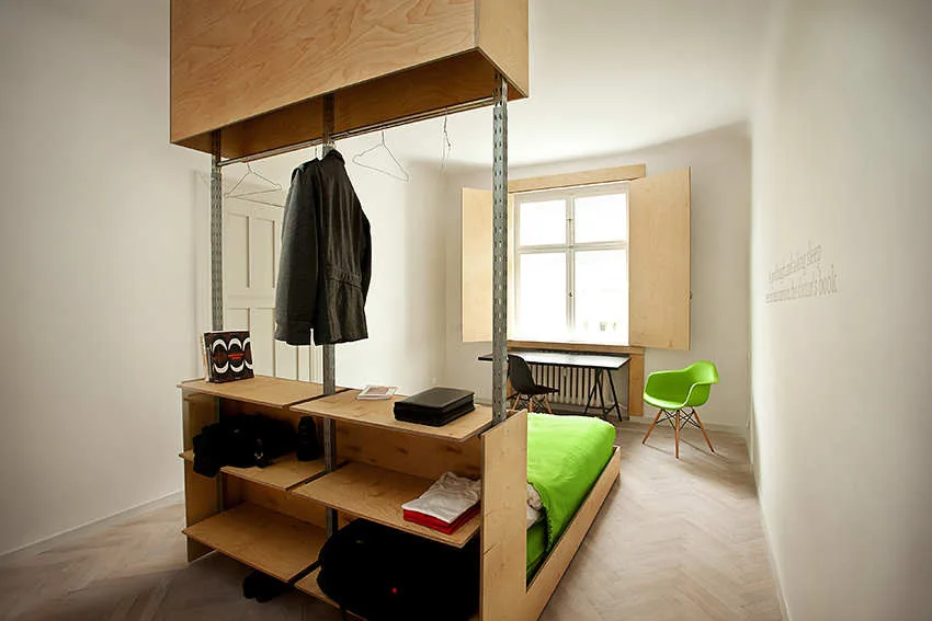 A minimalist bedroom features a custom plywood room divider with storage shelves, a green bed, and light herringbone floors.