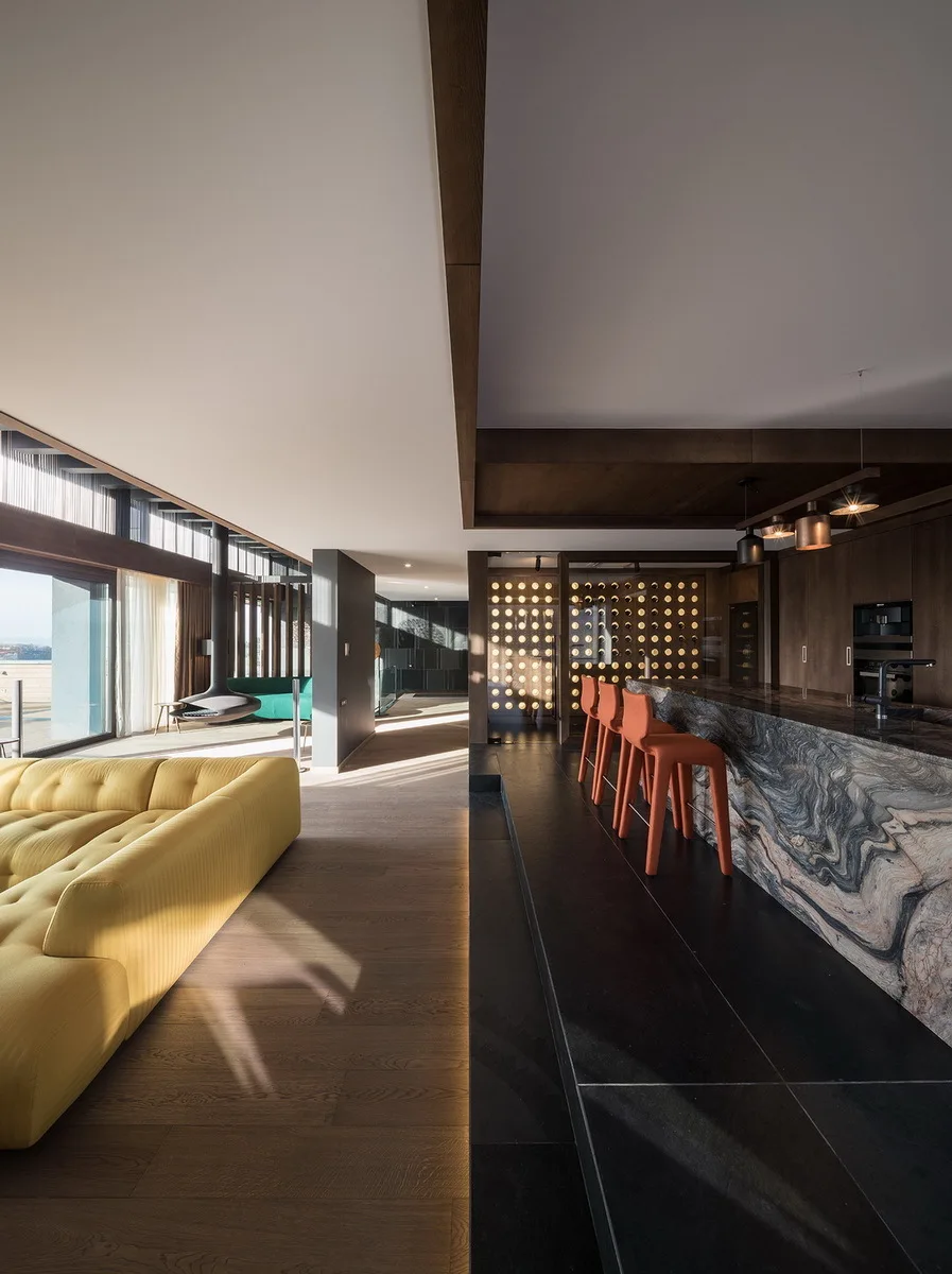 A modern open-plan kitchen and living room featuring a yellow tufted sofa, a heavily veined marble island with orange stools, and dark wood cabinetry.