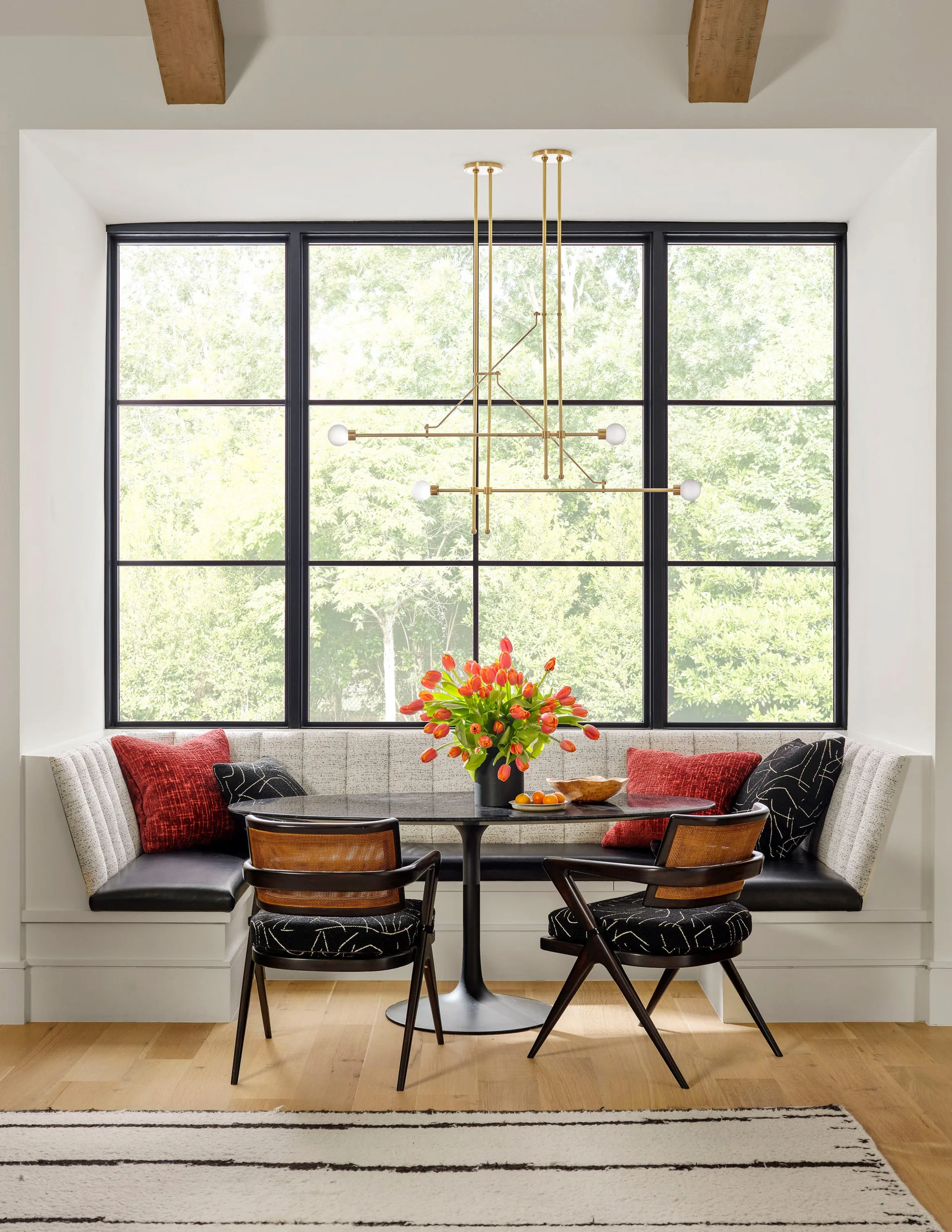 A modern breakfast nook features a built-in u-shaped bench, a black pedestal table, two cane-back chairs, and a large grid window overlooking trees.