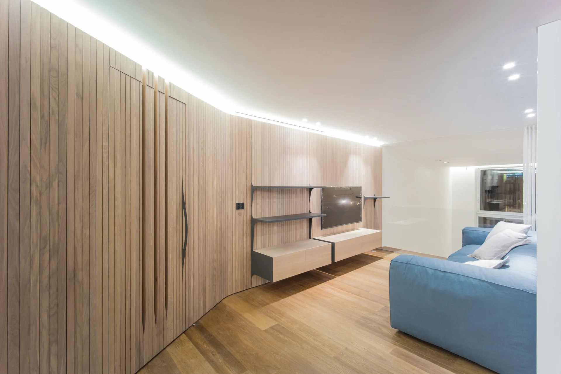 A contemporary room features a curved vertical-slat wood wall with a built-in television and storage shelves opposite a blue sofa.