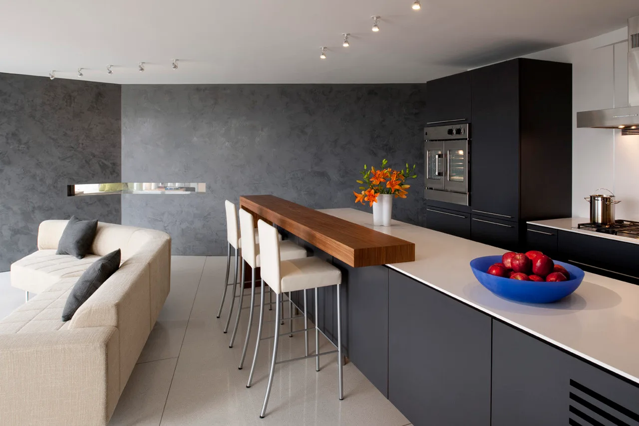 A modern kitchen featuring a gray island with a raised wood countertop, white bar stools, dark gray cabinets, and an adjacent light-colored sofa.