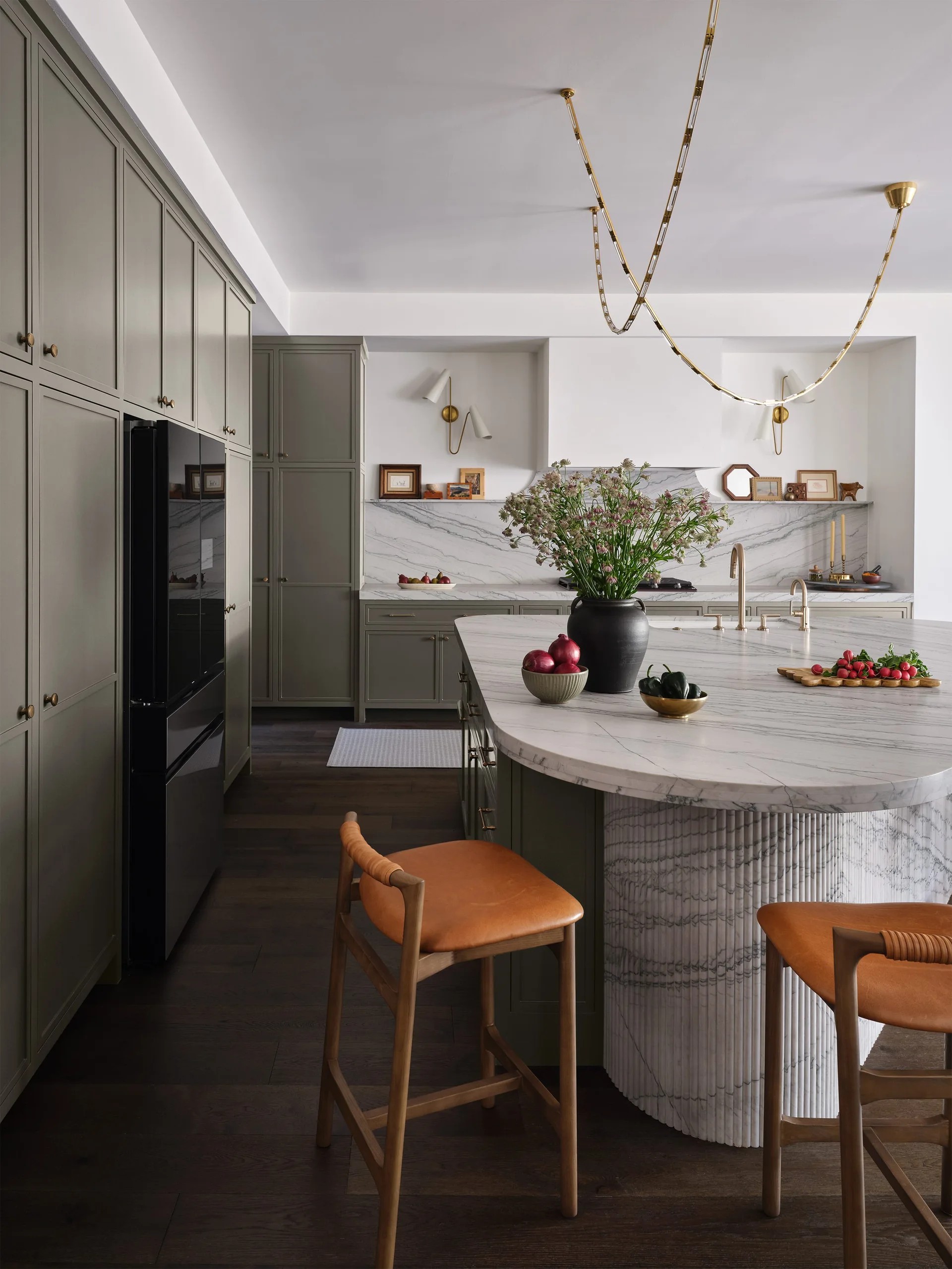 A kitchen features sage green cabinetry, a curved marble-topped island with a fluted base, leather bar stools, and a gold chain pendant light.