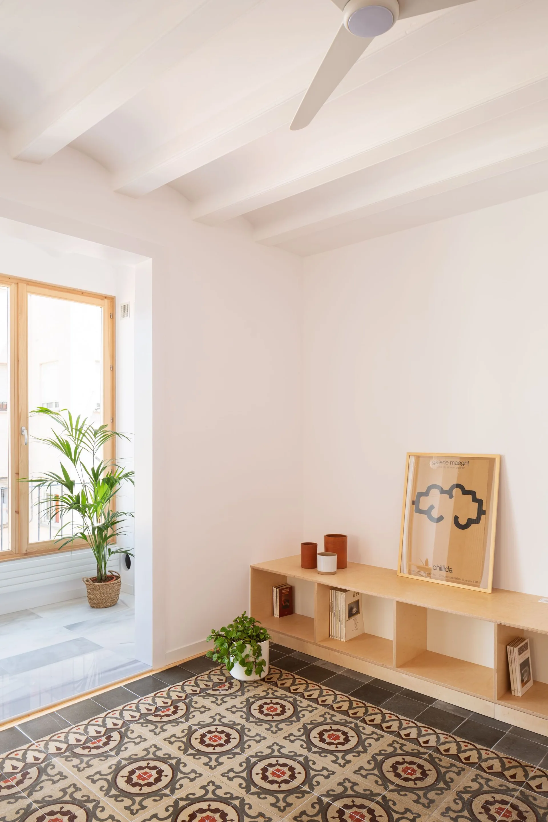 A minimalist room features a low wooden cubby shelf on patterned ceramic floor tiles near a white ceiling with exposed beams.
