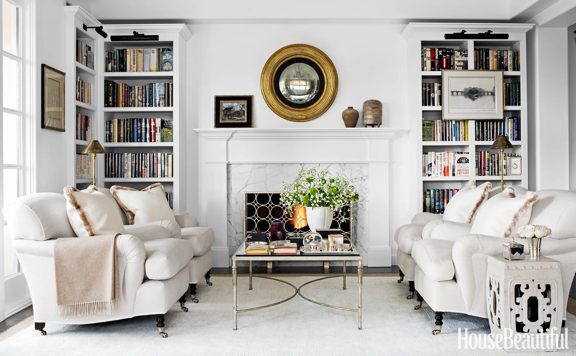 A symmetrical white living room features a marble fireplace flanked by tall built-in bookshelves, two off-white sofas, and a gold round mirror.