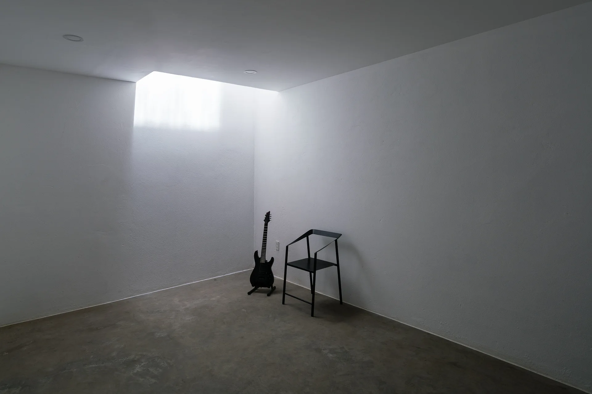 A minimalist white room with a concrete floor, featuring a black electric guitar and a modern black metal chair under an overhead skylight.