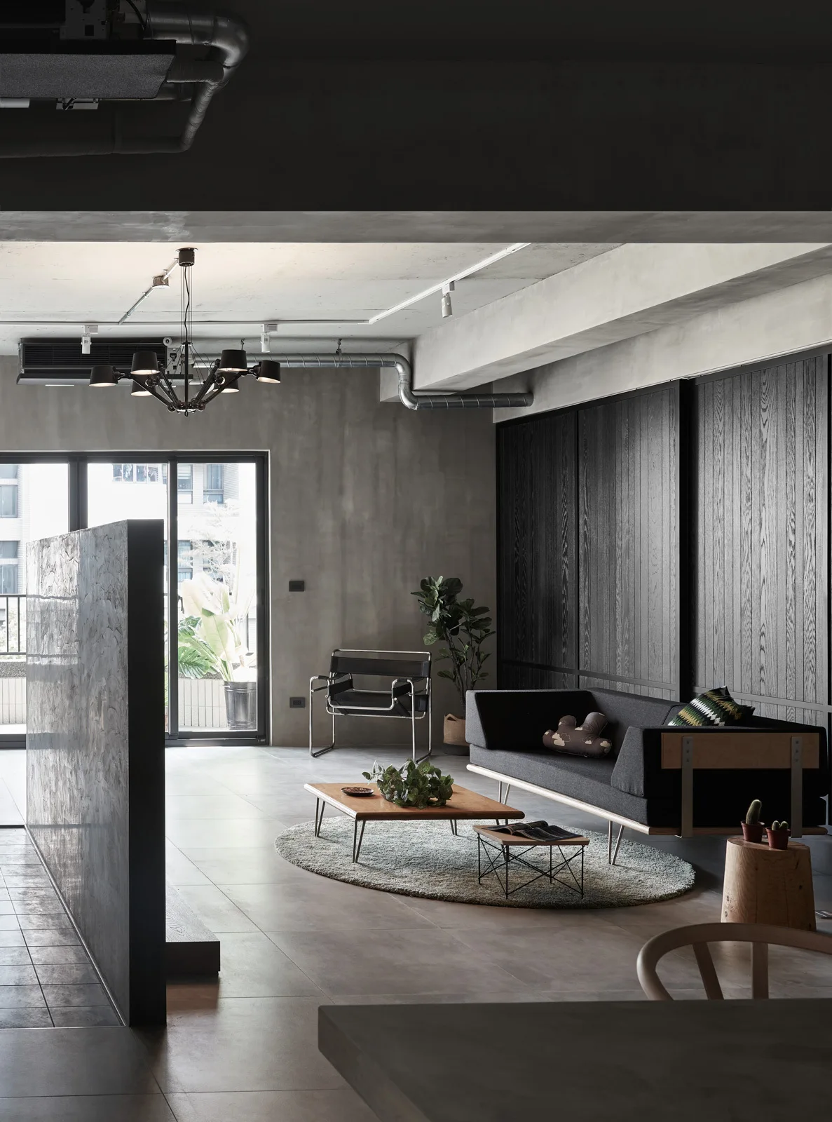 An industrial style living room features a charcoal grey sofa, a low wooden coffee table on a round rug, concrete walls, and dark wood vertical paneling.