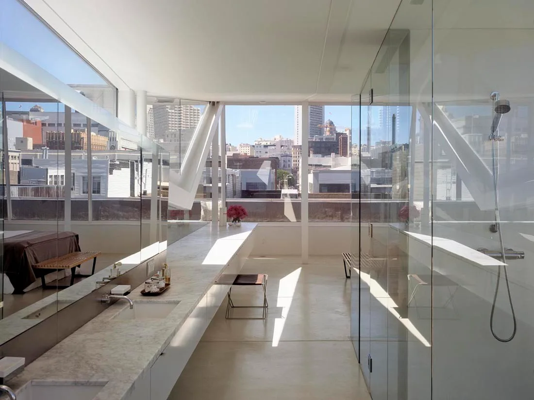 A minimalist modern bathroom features a long marble vanity with sinks, a glass-enclosed shower, and large windows overlooking a city skyline.