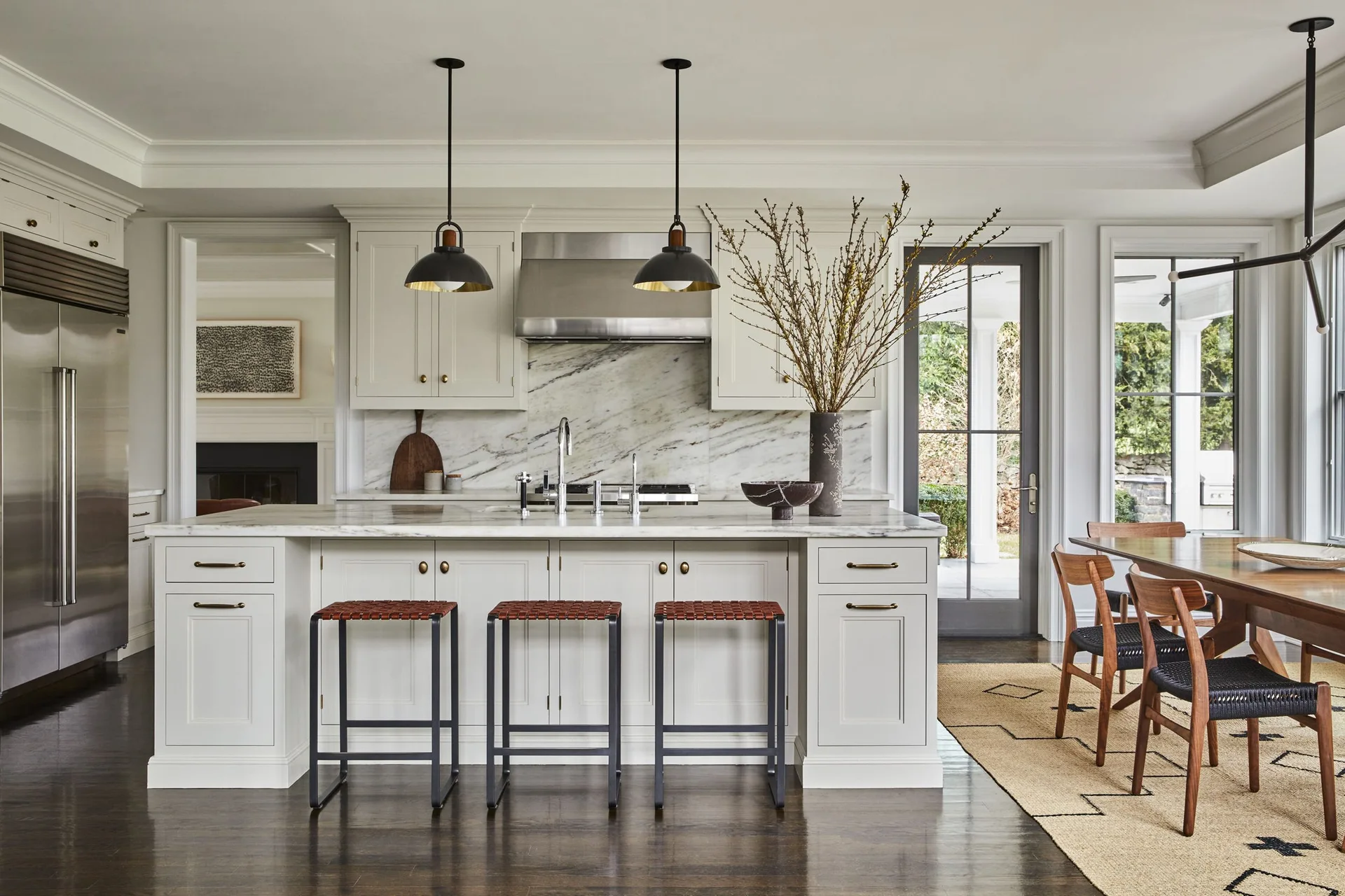 A transitional kitchen features a marble-topped island with woven leather stools, black dome pendant lights, and off-white cabinetry.
