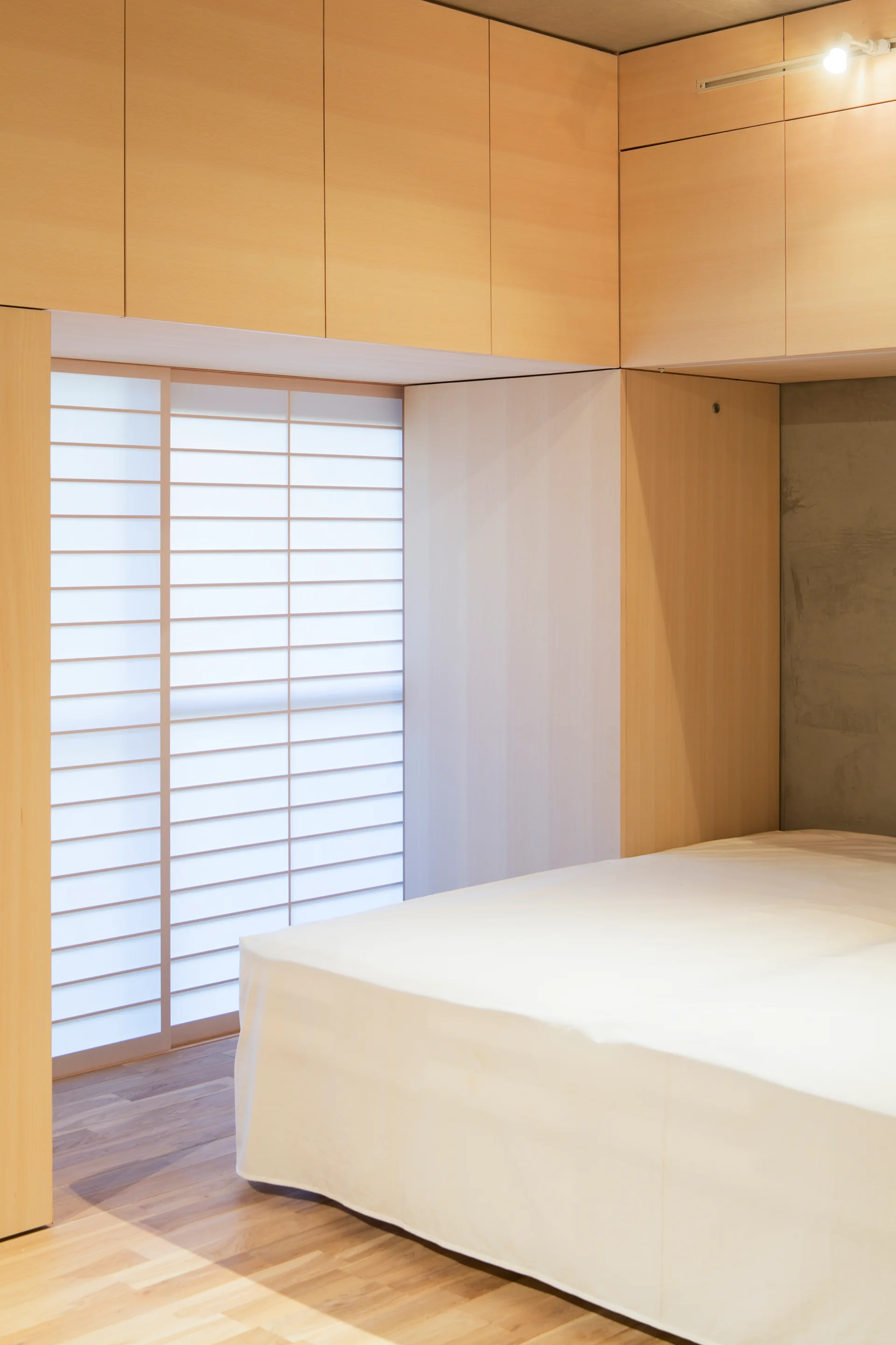 A minimalist bedroom features a bed with white linens, built-in light wood cabinetry, an exposed concrete wall, and a sliding shoji screen.