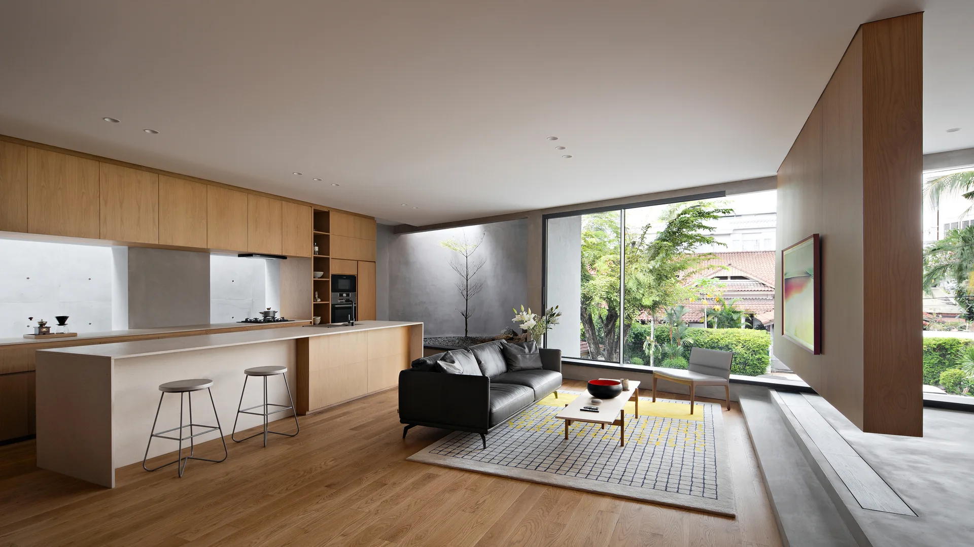 A modern open-plan kitchen and living room features light wood cabinetry, a long island, a black leather sofa on a grid rug, and a large window.