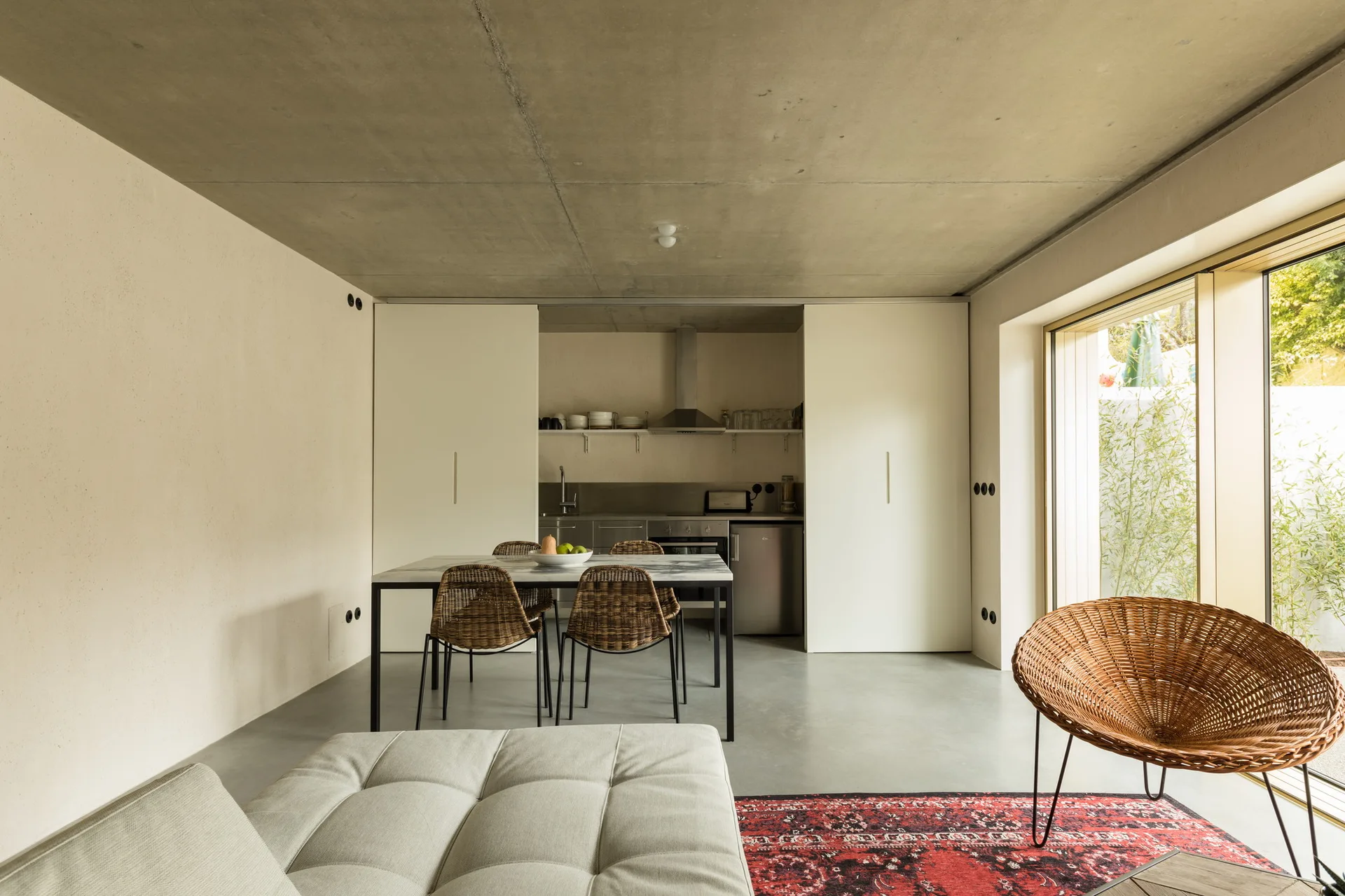An open-plan dining and living space features an exposed concrete ceiling, a dining table with wicker chairs, a red patterned rug, and a recessed kitchen.