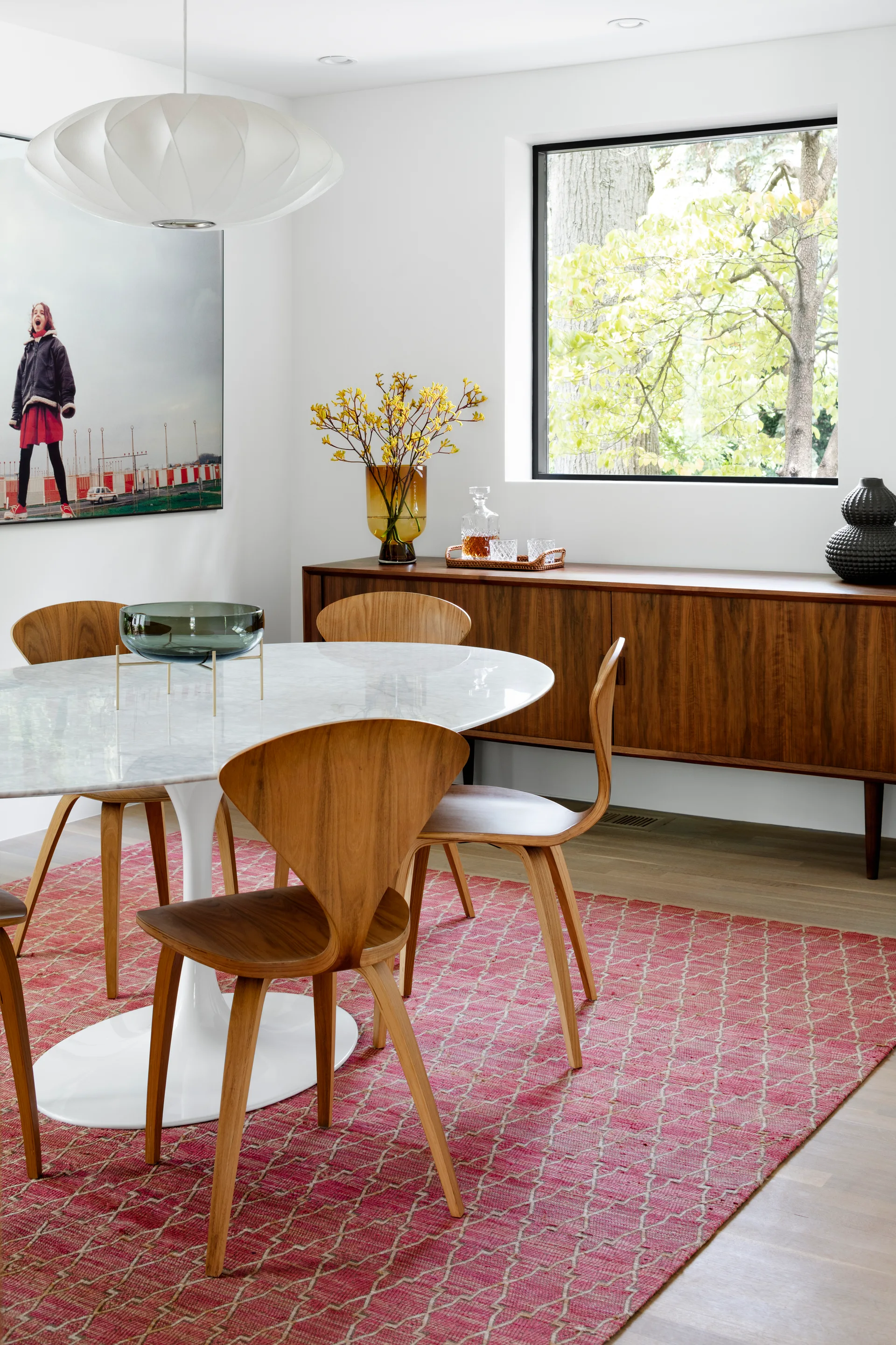 An oval white marble dining table is surrounded by curved wood veneer chairs on a pink patterned rug near a walnut sideboard.