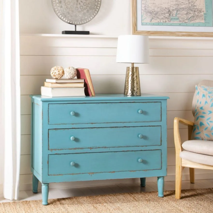 A distressed light blue dresser stands on a woven jute rug against a white shiplap wall with a gold lamp and books on top.