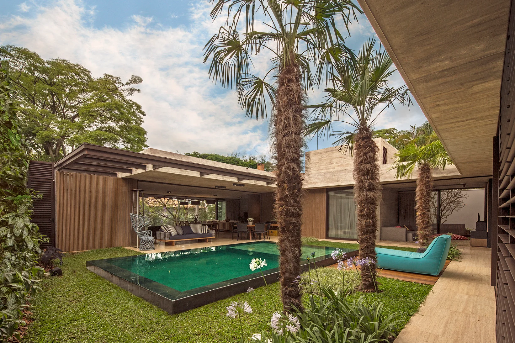 A modern backyard patio features an in-ground swimming pool, palm trees, a concrete pavilion, and a bright blue lounge chair.