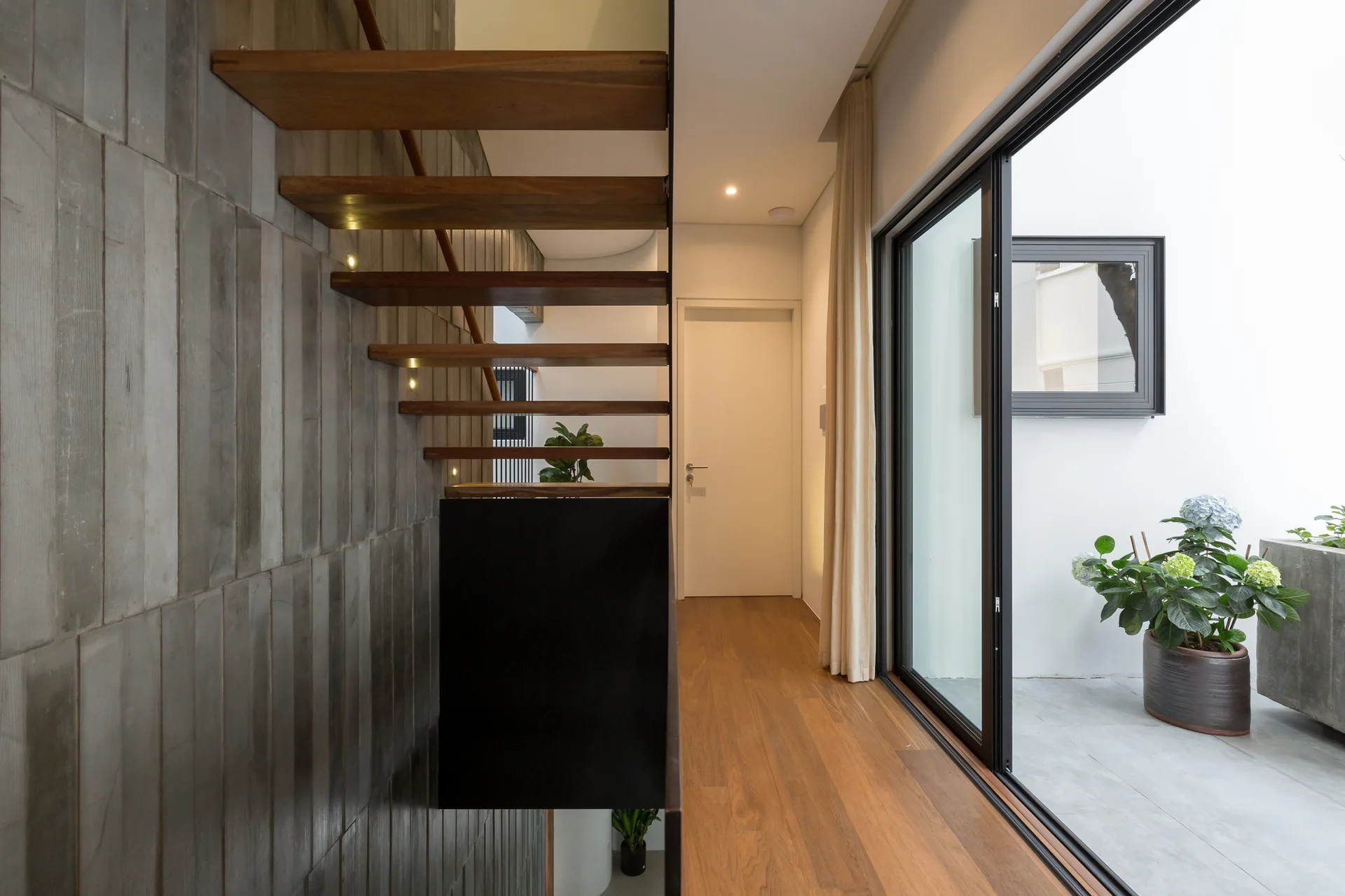 A modern hallway features floating wooden stairs against a textured concrete wall, next to a large sliding glass door leading to a patio.