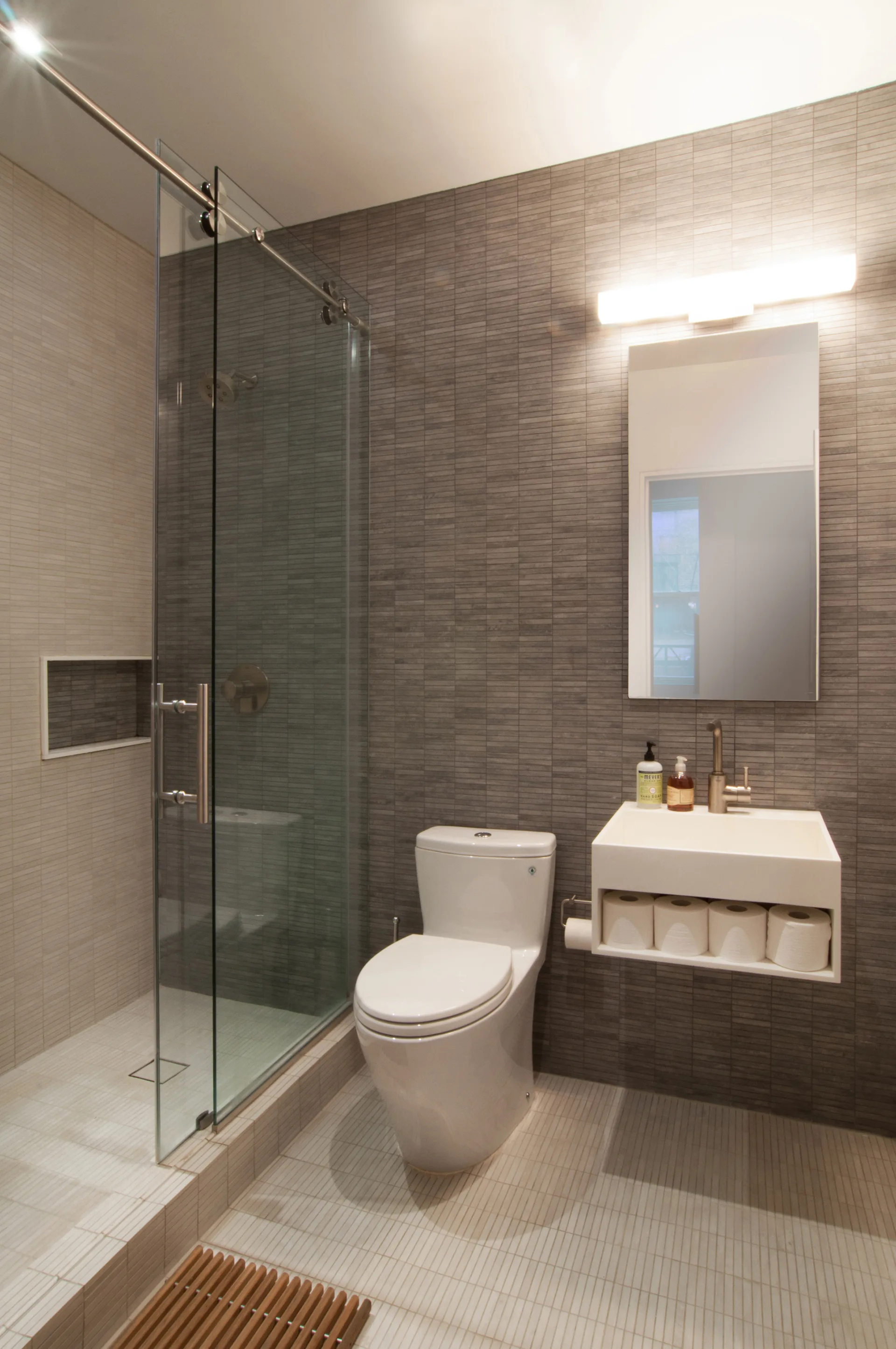 A modern bathroom featuring grey horizontal tile walls, a white toilet, a floating rectangular sink with open storage, and a glass-enclosed shower.