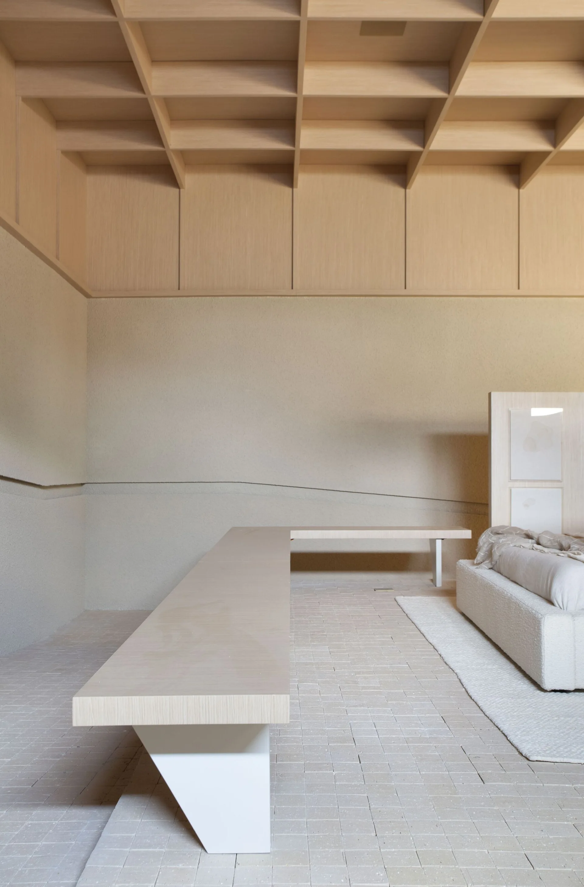 A minimalist bedroom features a light wood coffered ceiling, textured beige walls, a low platform bed, and a long wood bench on a tiled floor.