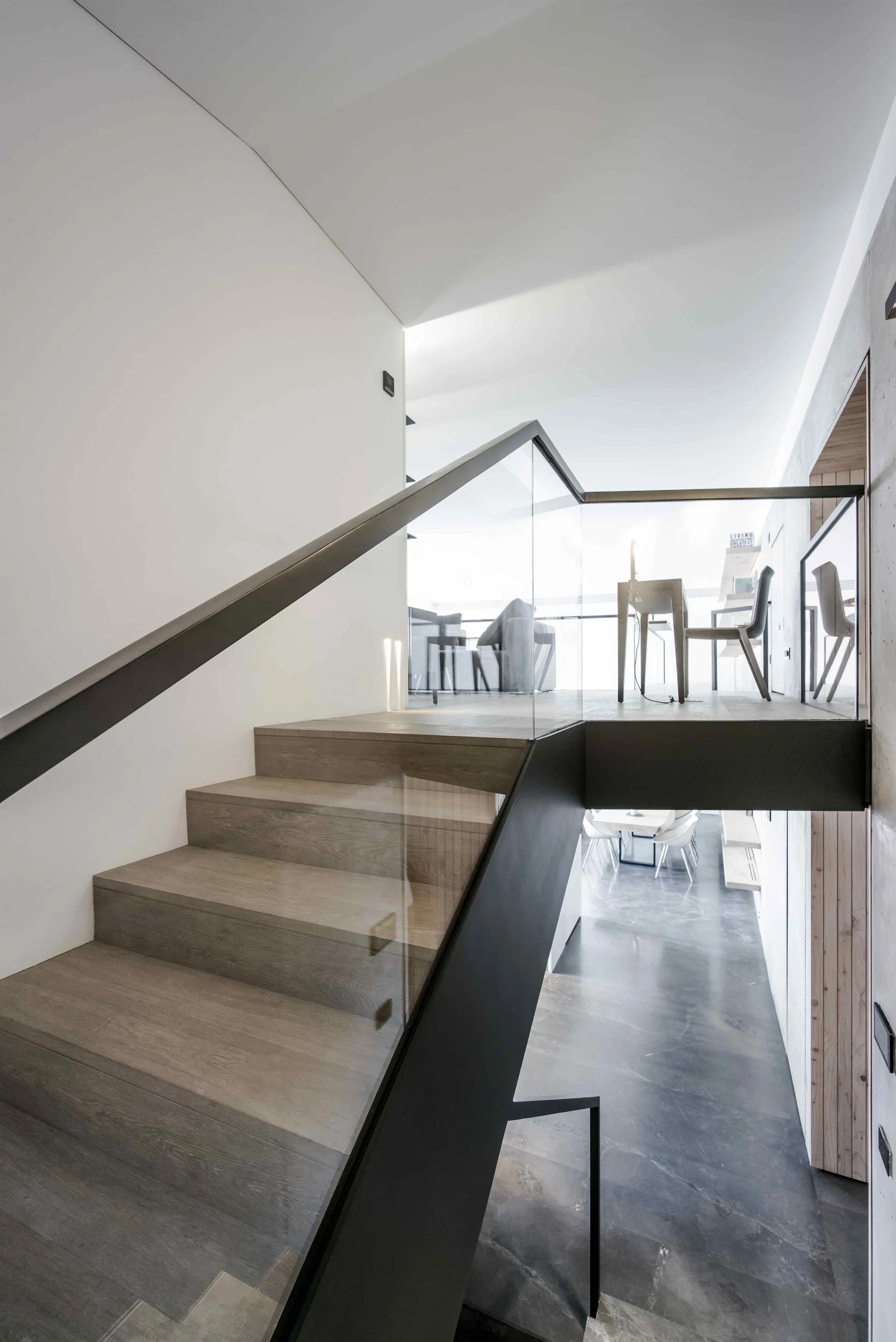 A modern staircase with light wood treads and a glass railing with black steel framing rises toward a bright upper landing with chairs and a table.