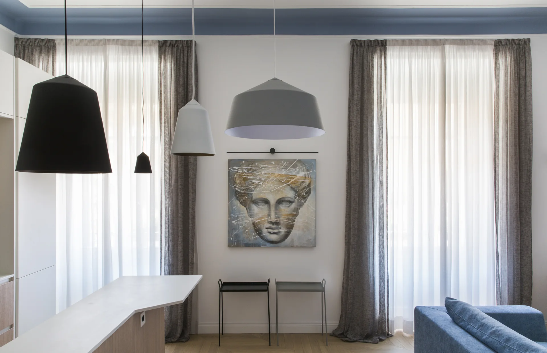 A contemporary room features hanging pendant lights, sheer grey curtains, a central face painting, and a blue painted border along the ceiling.
