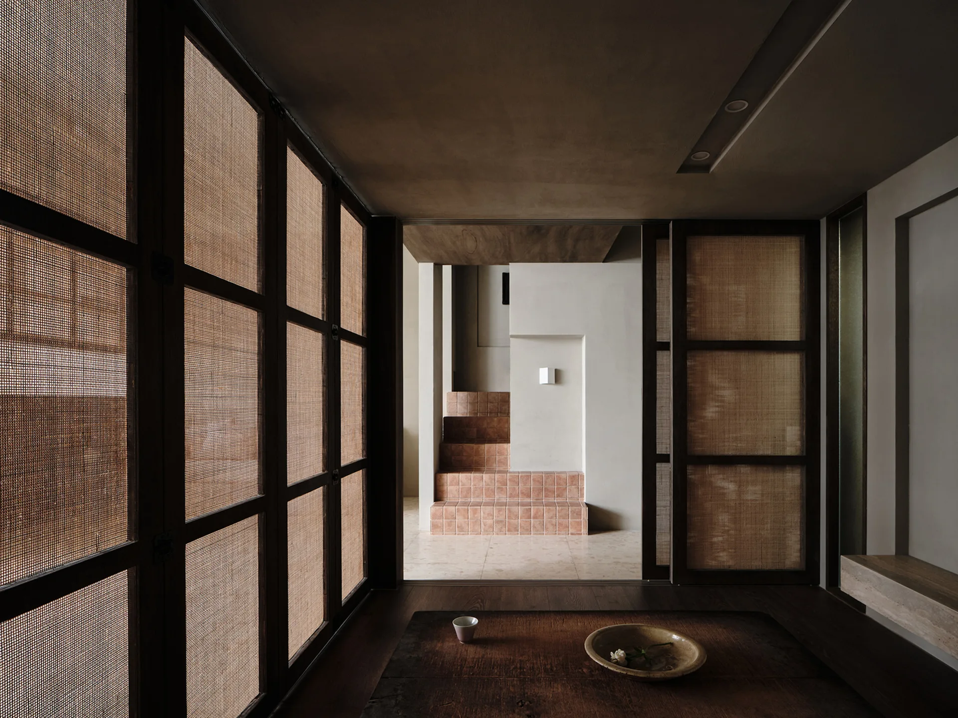 A minimalist room features woven rattan sliding doors that look onto a passage with a small terracotta tiled staircase.