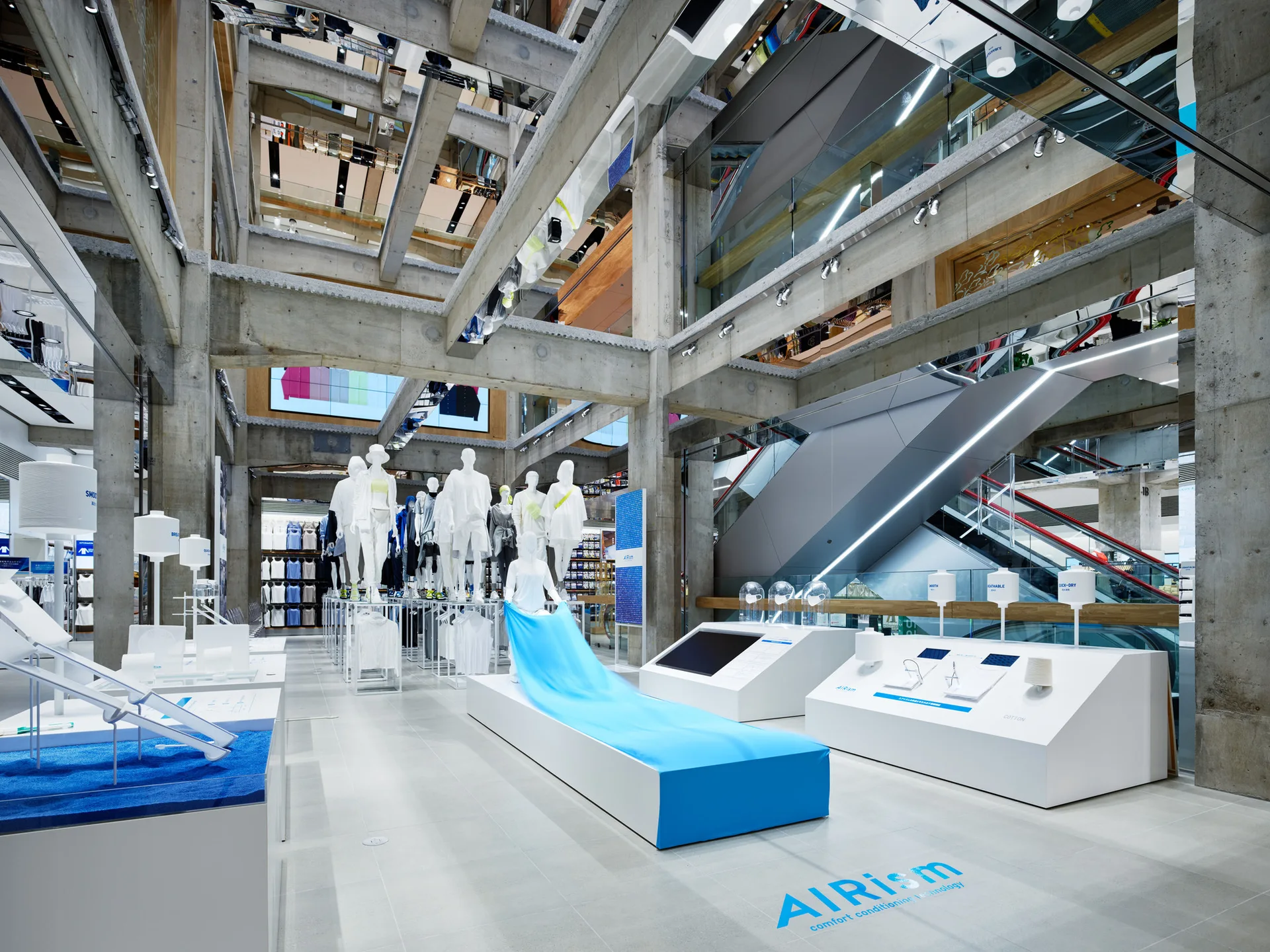 A spacious retail interior featuring exposed concrete columns and ceiling beams, minimalist white display stands, and mannequins.
