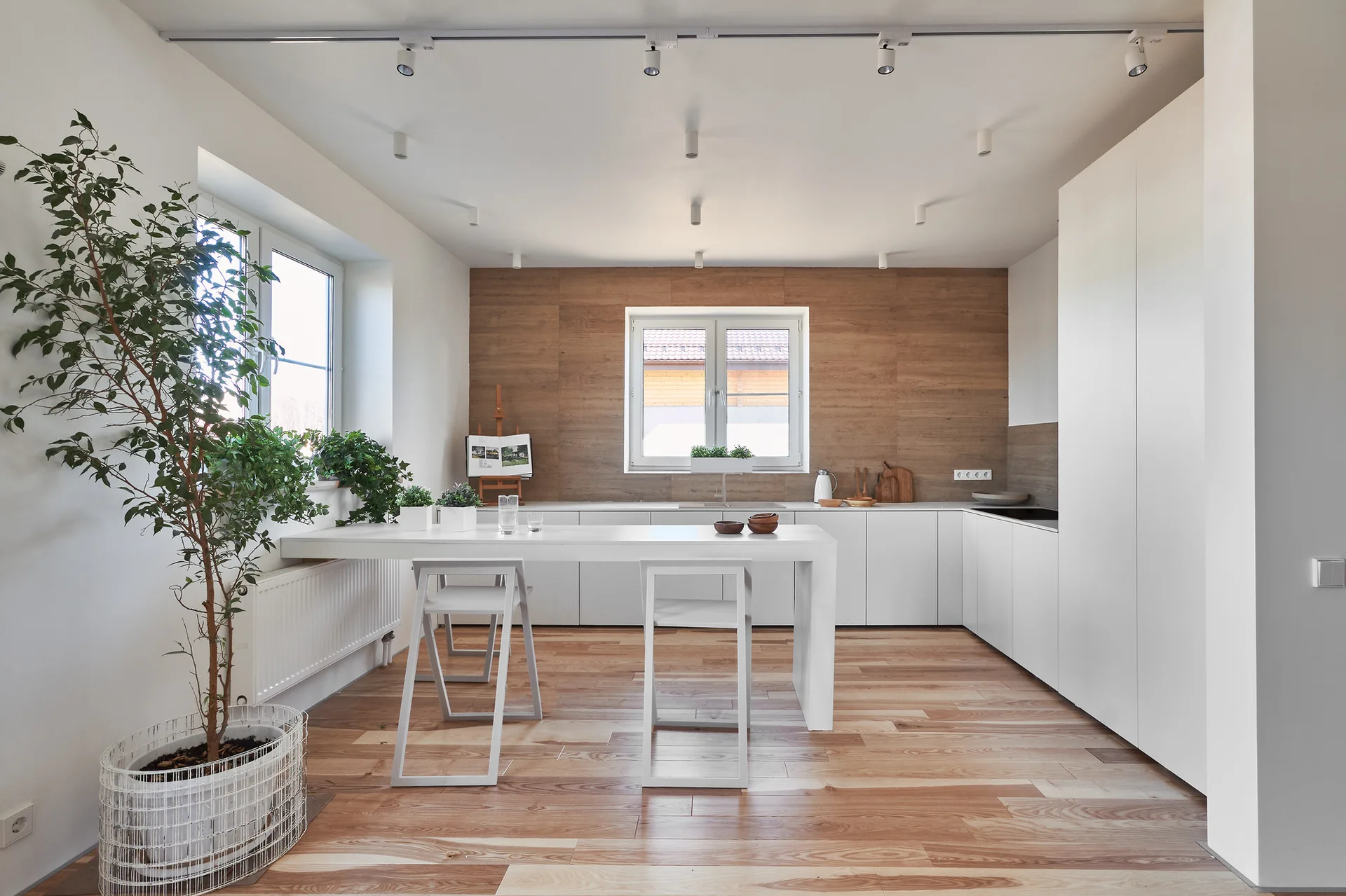 A minimalist white kitchen features flat-front cabinets, a matching dining table, light wood flooring, and a warm wood-paneled accent wall.