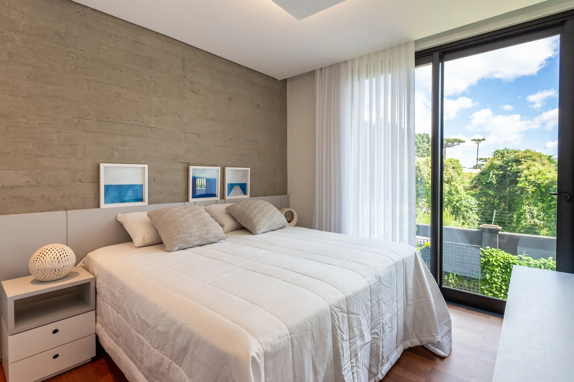 A minimalist bedroom featuring a board-formed concrete wall behind a bed with white bedding next to a large sliding glass door with sheer curtains.