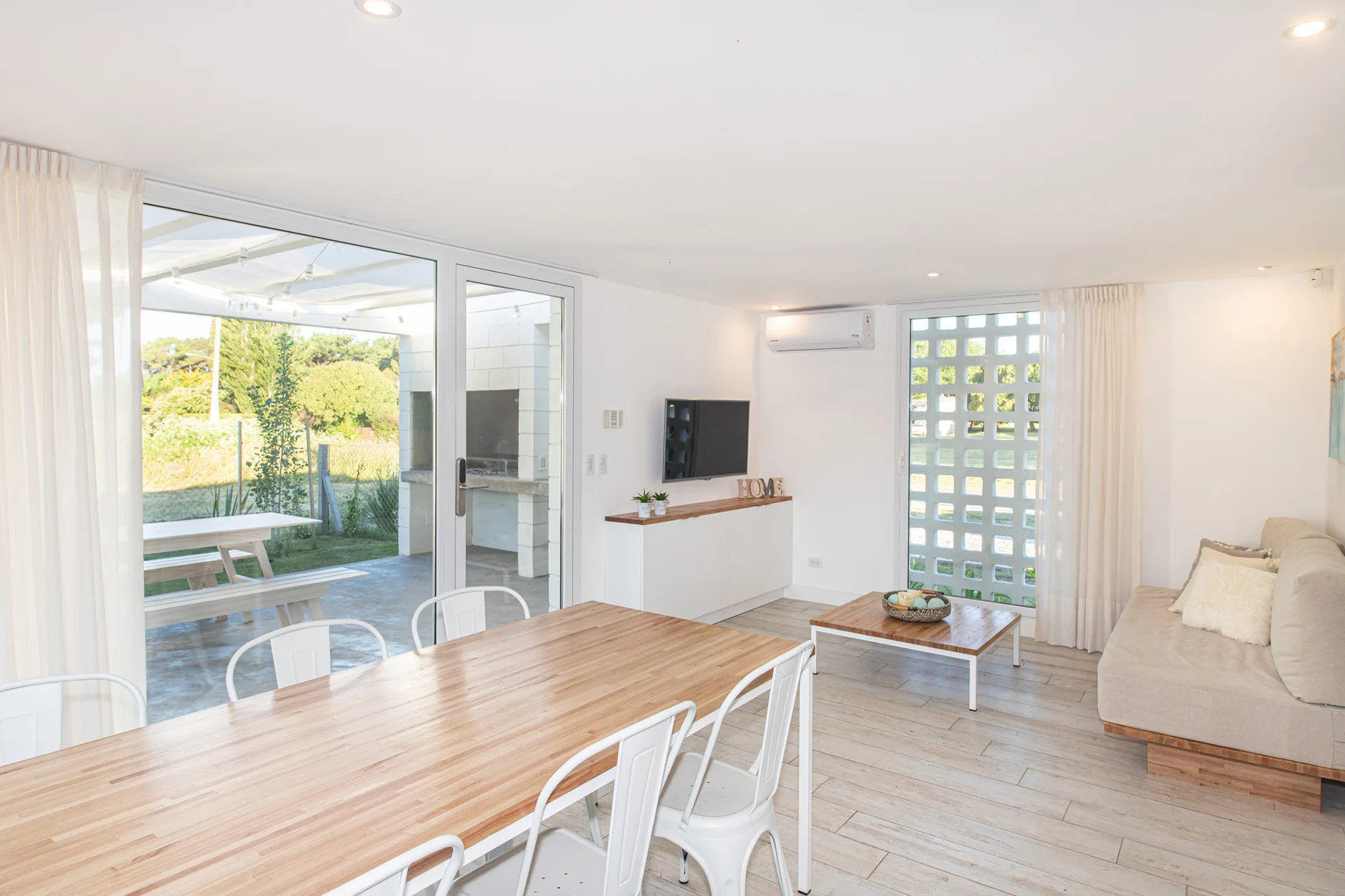 A bright open-plan dining and living space features a light wood table with white metal chairs and glass doors opening to a patio.