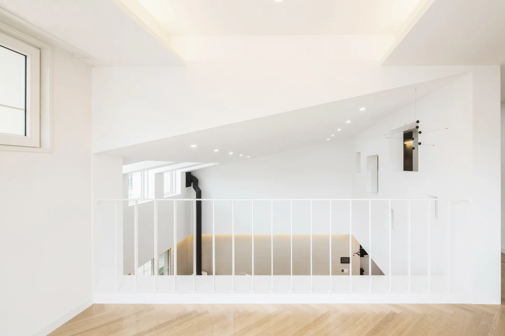 A minimalist loft landing featuring a thin white metal railing, light-toned herringbone wood floors, and an angled ceiling with recessed lighting.