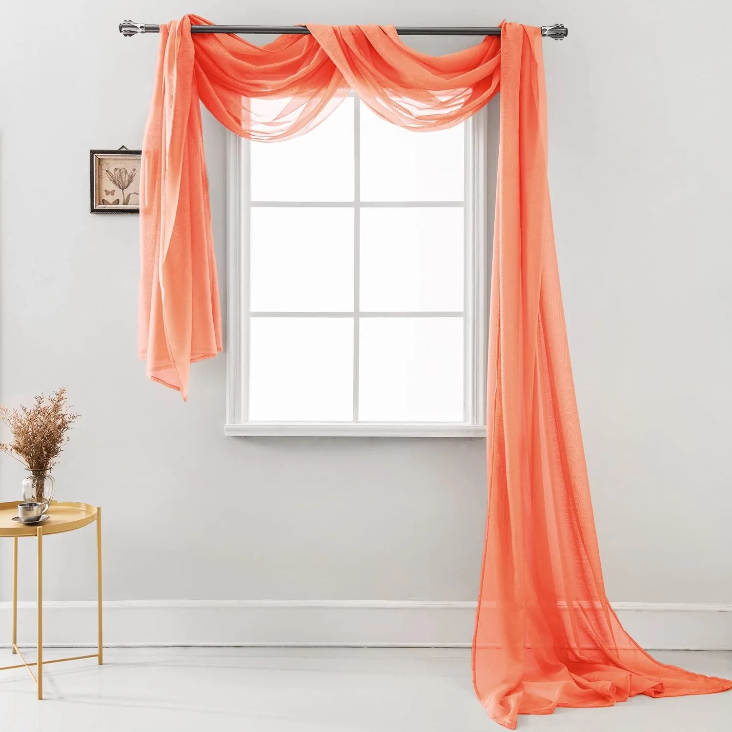 A sheer coral fabric scarf curtain is draped asymmetrically over a black metal rod above a white window in a bright room.