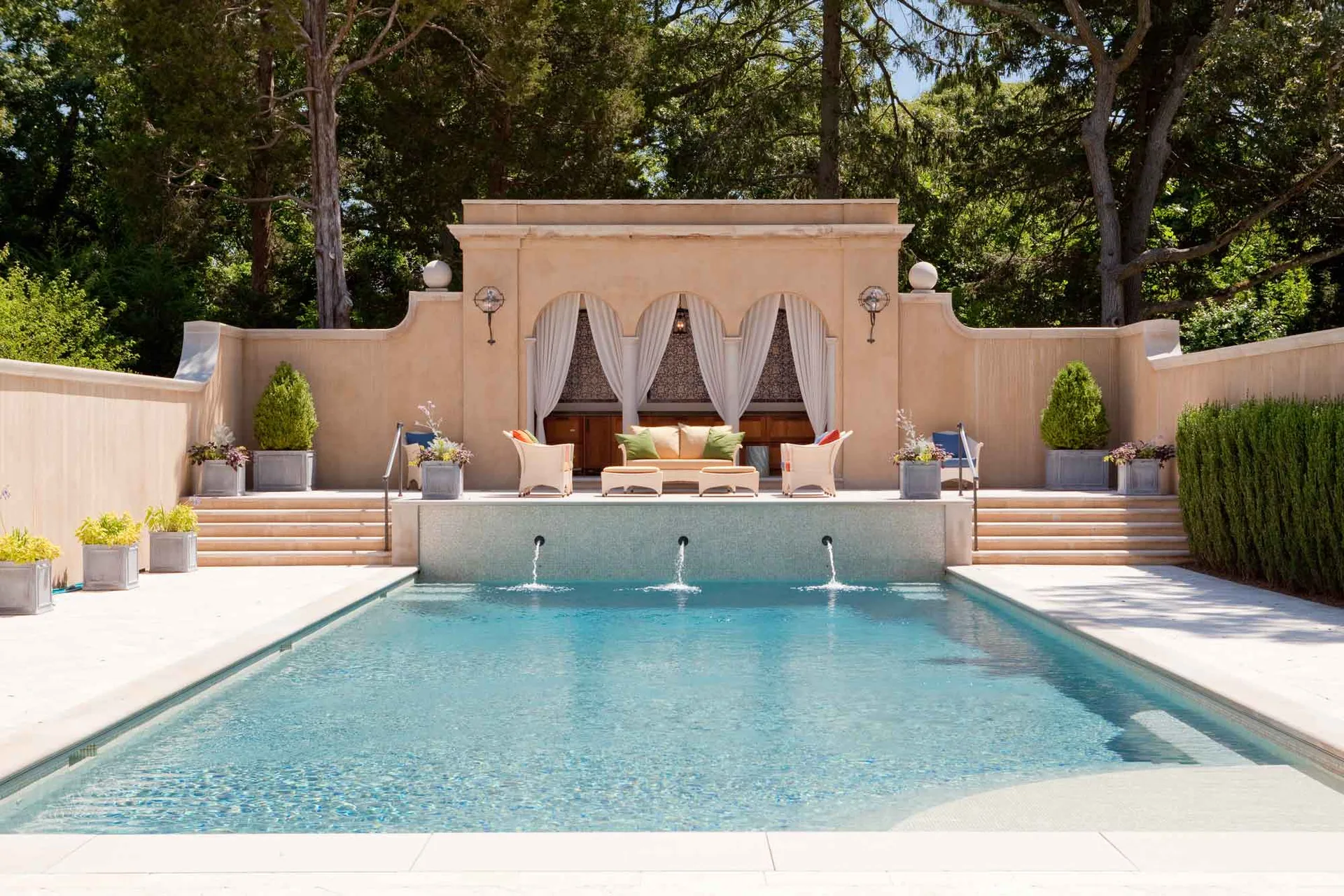 A rectangular swimming pool with three water fountains spouting from a raised beige stone patio featuring a symmetrical arched cabana and outdoor seating.