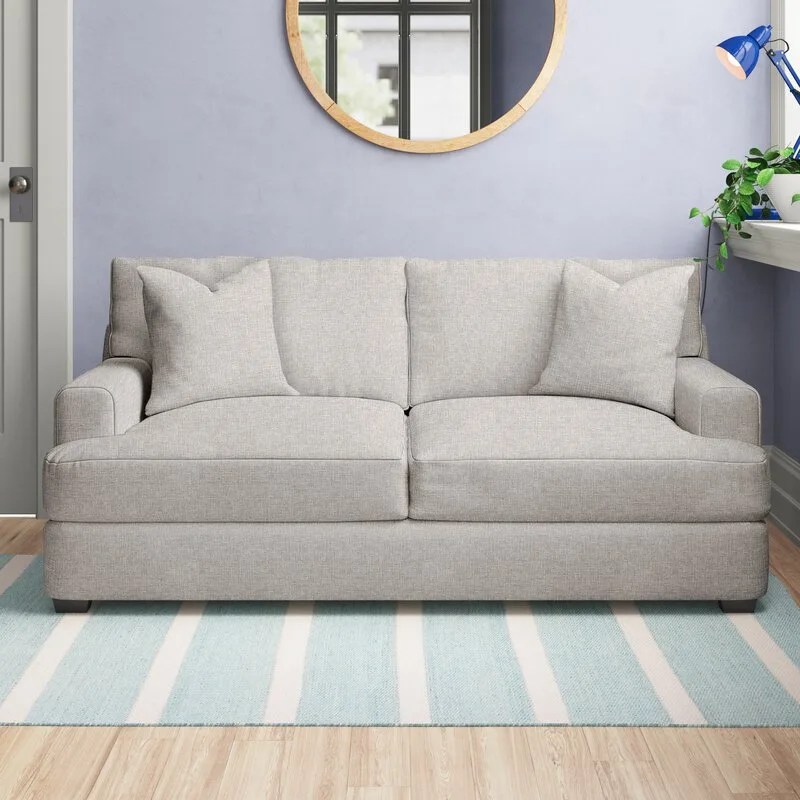 A light grey upholstered sofa sits on a blue and white striped rug in front of a periwinkle wall with a round wood-framed mirror.