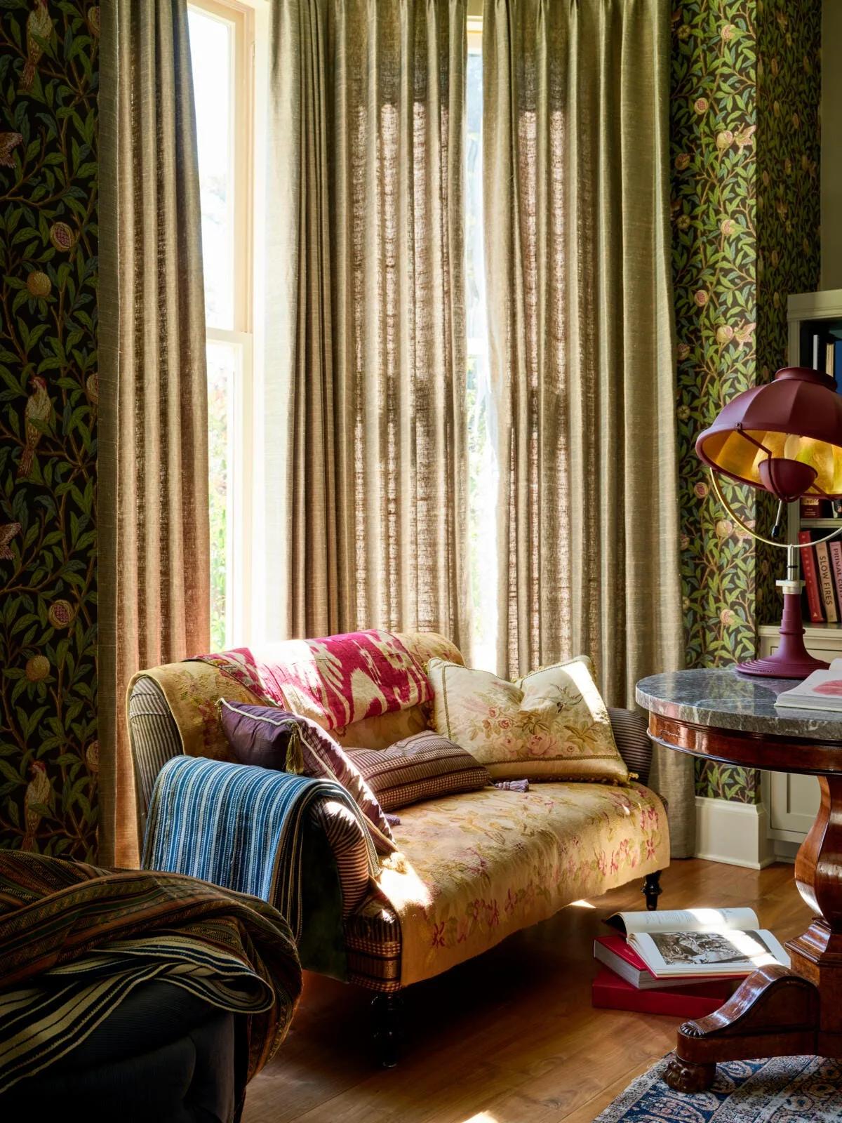 An upholstered floral settee sits in front of tall brown curtains and botanical print wallpaper in a sunlit room.