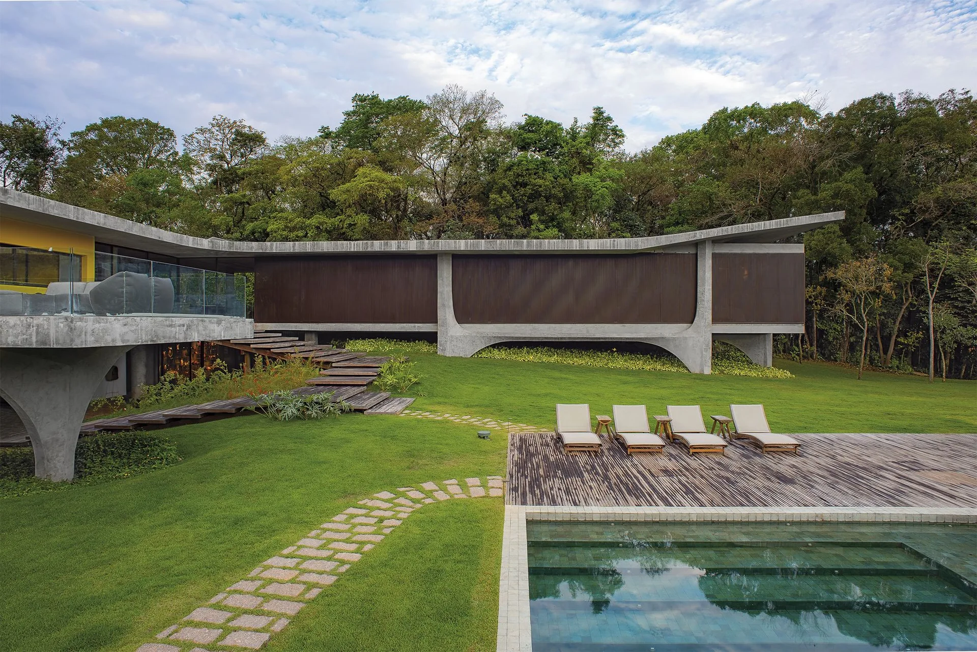 A modern concrete home stands above a grassy lawn next to a rectangular swimming pool with four lounge chairs on a wooden deck.