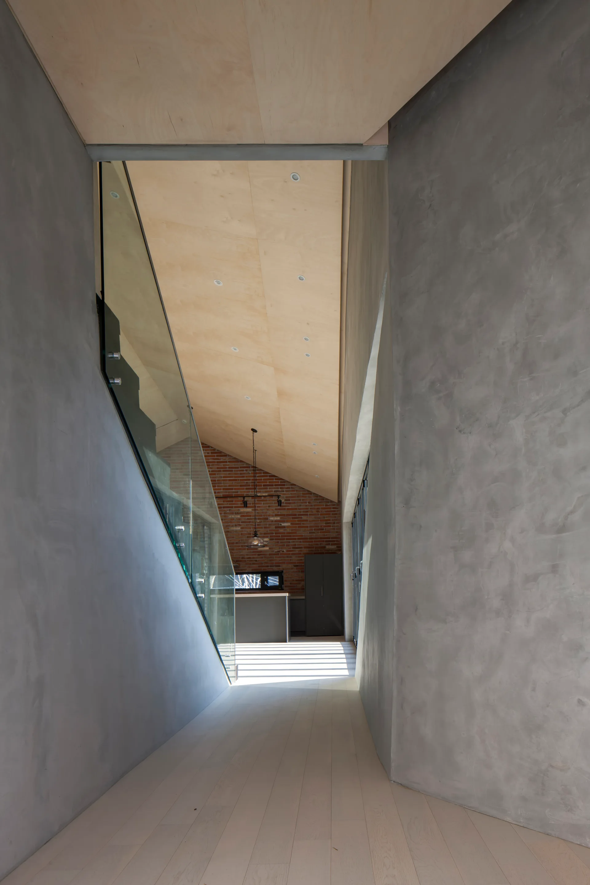 A modern hallway with matte grey concrete walls, a light plywood ceiling, and a glass staircase balustrade leading toward a kitchen with a brick wall.