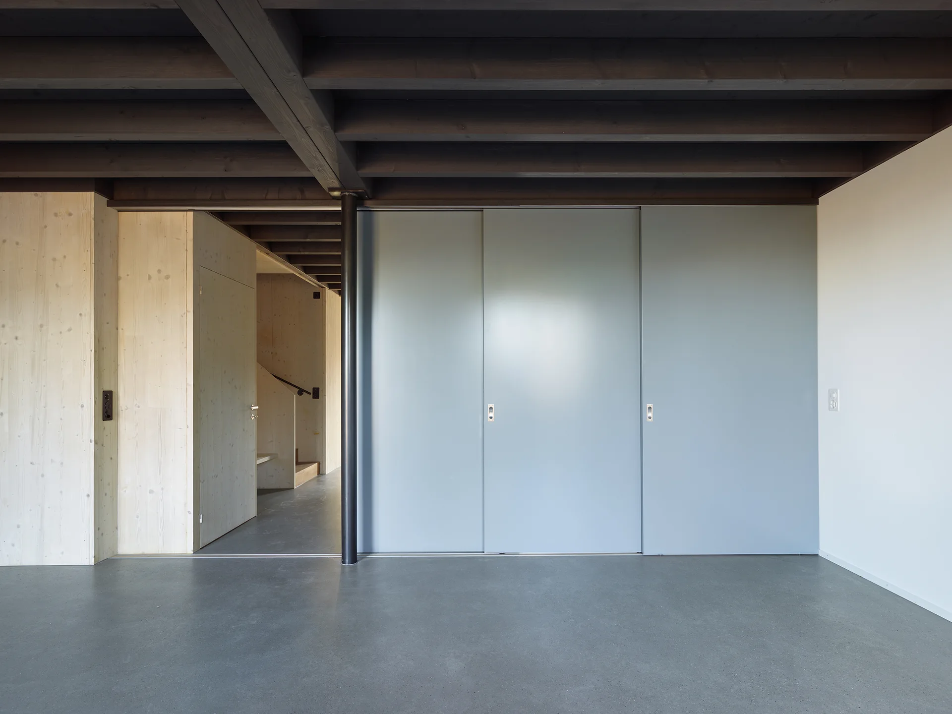 A modern room features dark exposed ceiling beams, light wood-clad walls, grey sliding doors, and a smooth grey concrete floor.